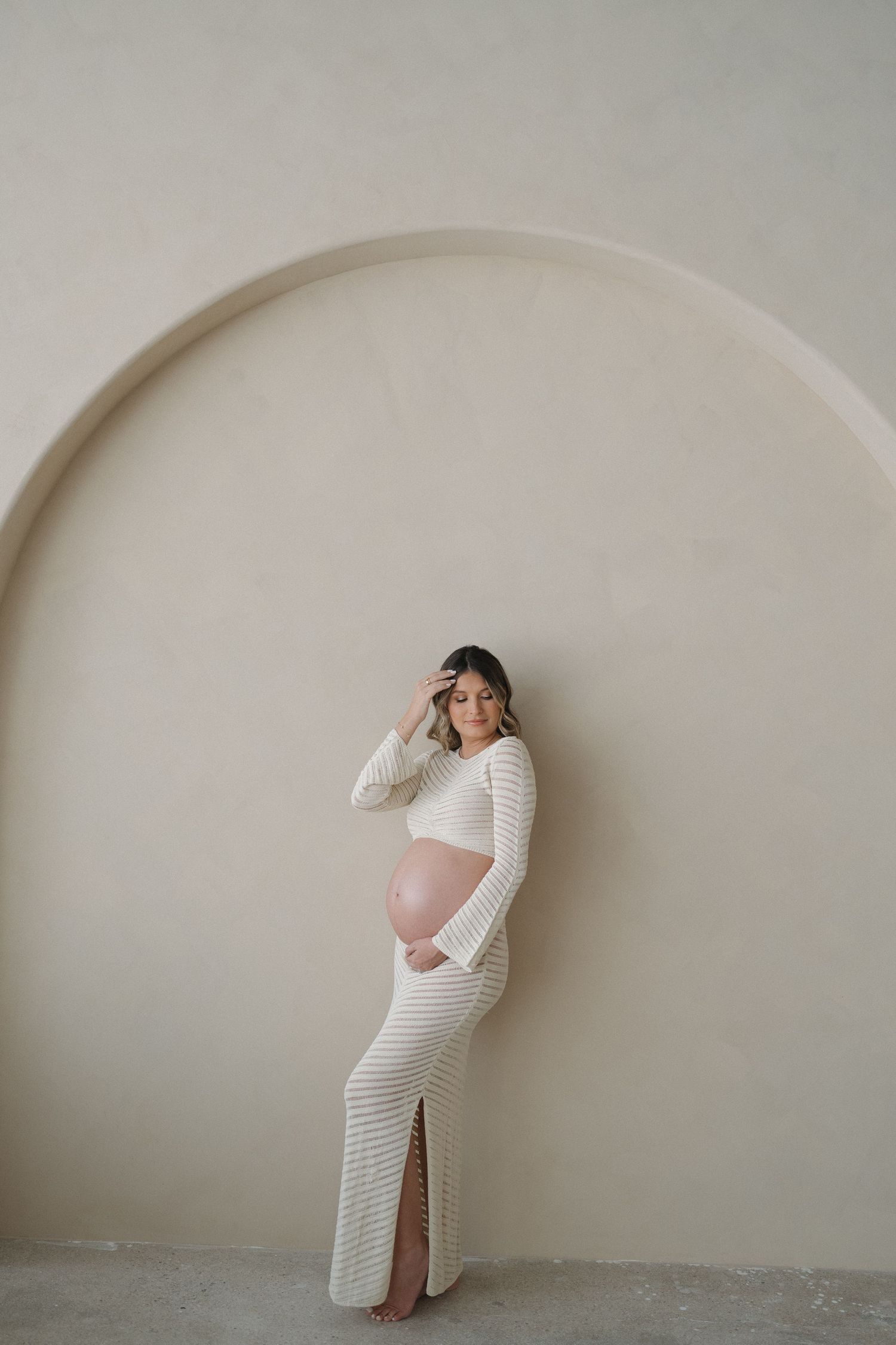 Expectant mother poses in white striped dress against neutral wall with curved archway.