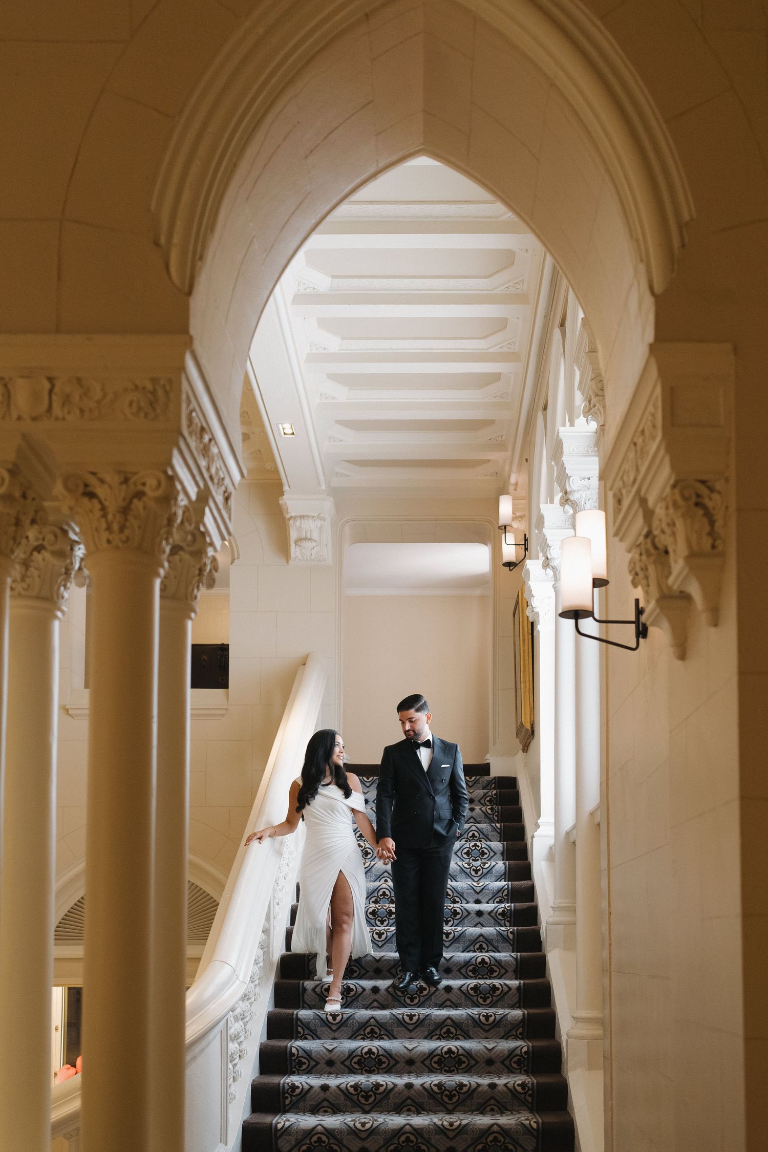 A wedding couple stands on ornate marble stairs beneath an elegant arched corridor.