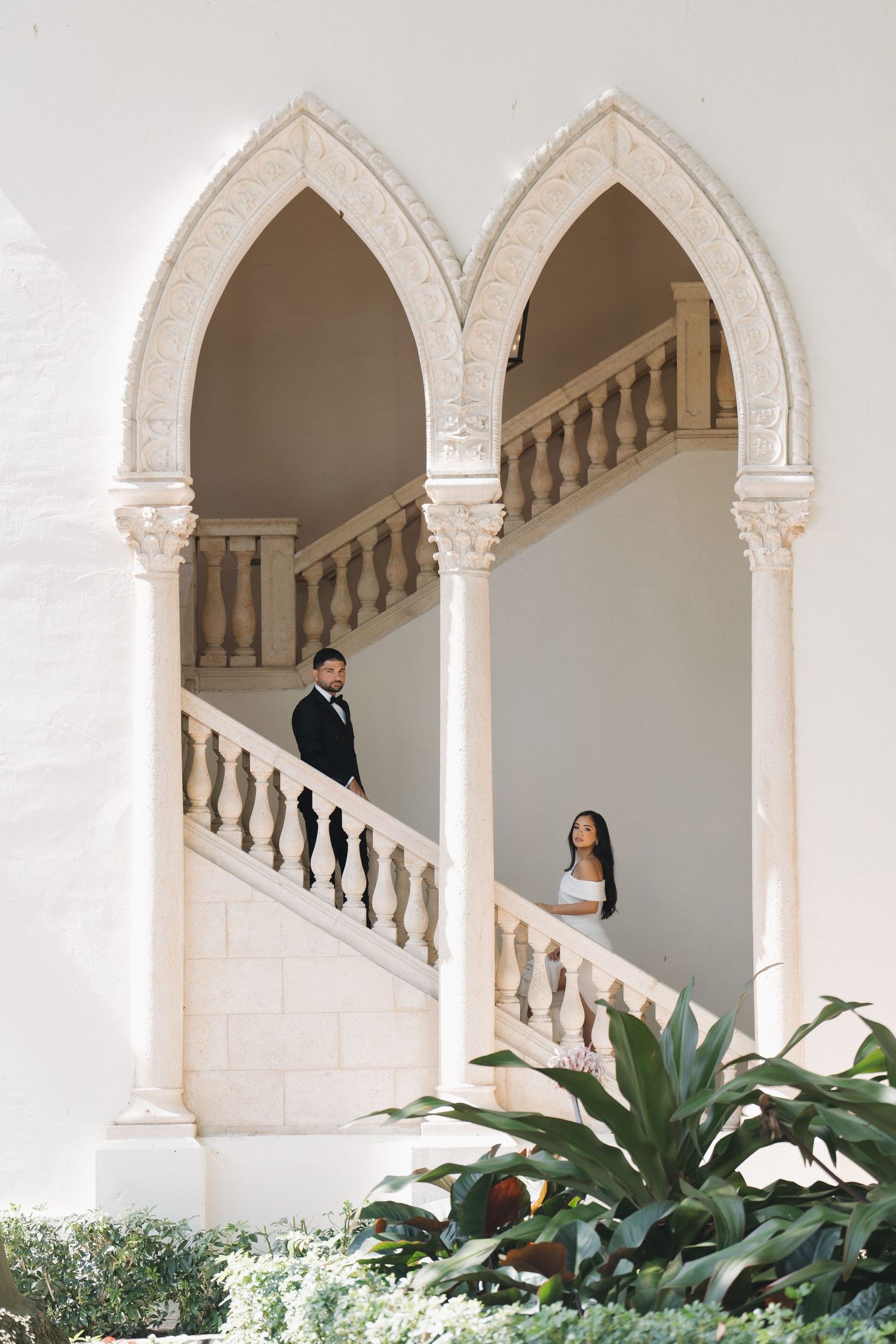 An elegant white stone archway with balustrades decorated with tropical greenery creates a romantic architectural scene.