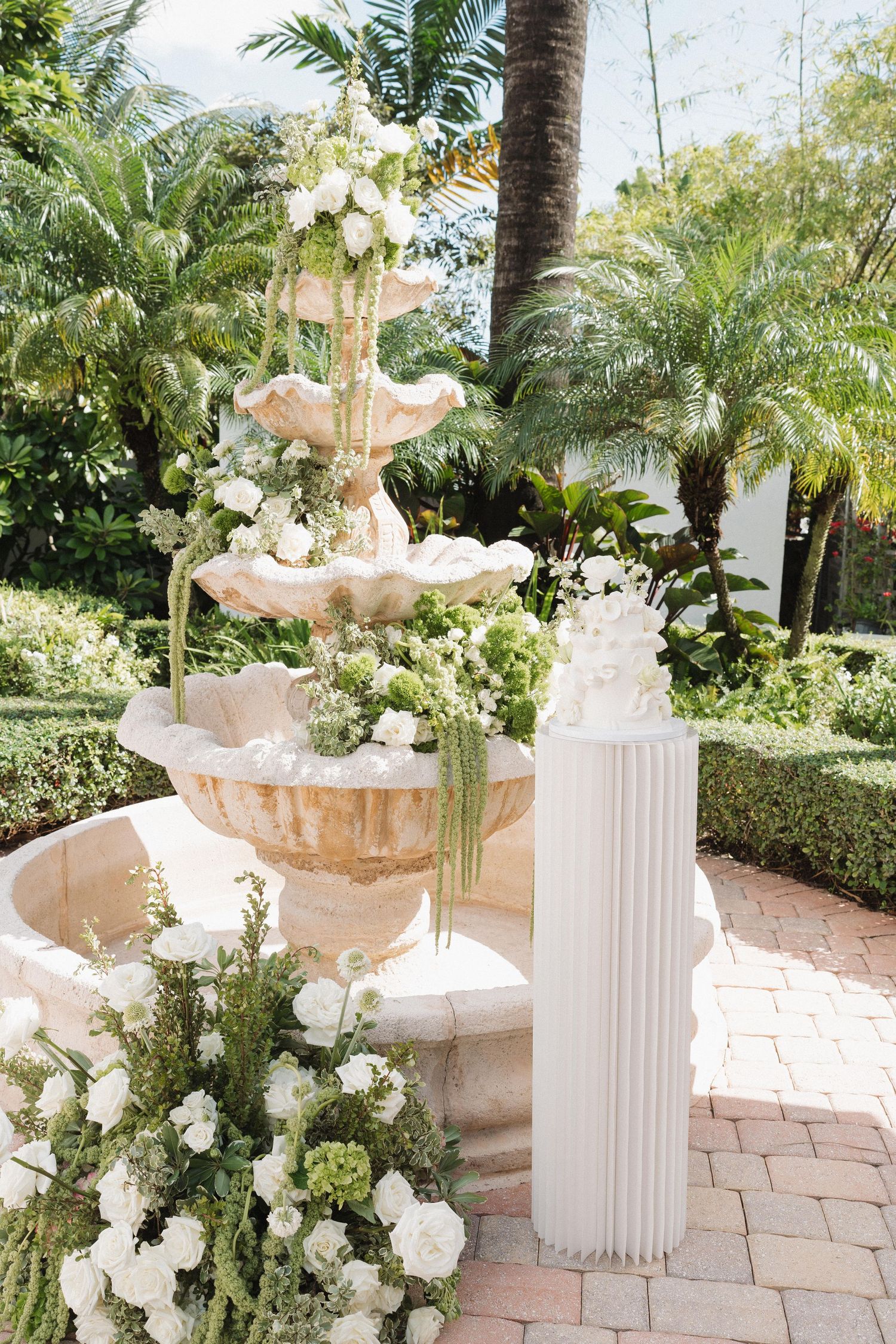 Elegant white floral arrangements in stone urns line a garden pathway with tropical palm trees.