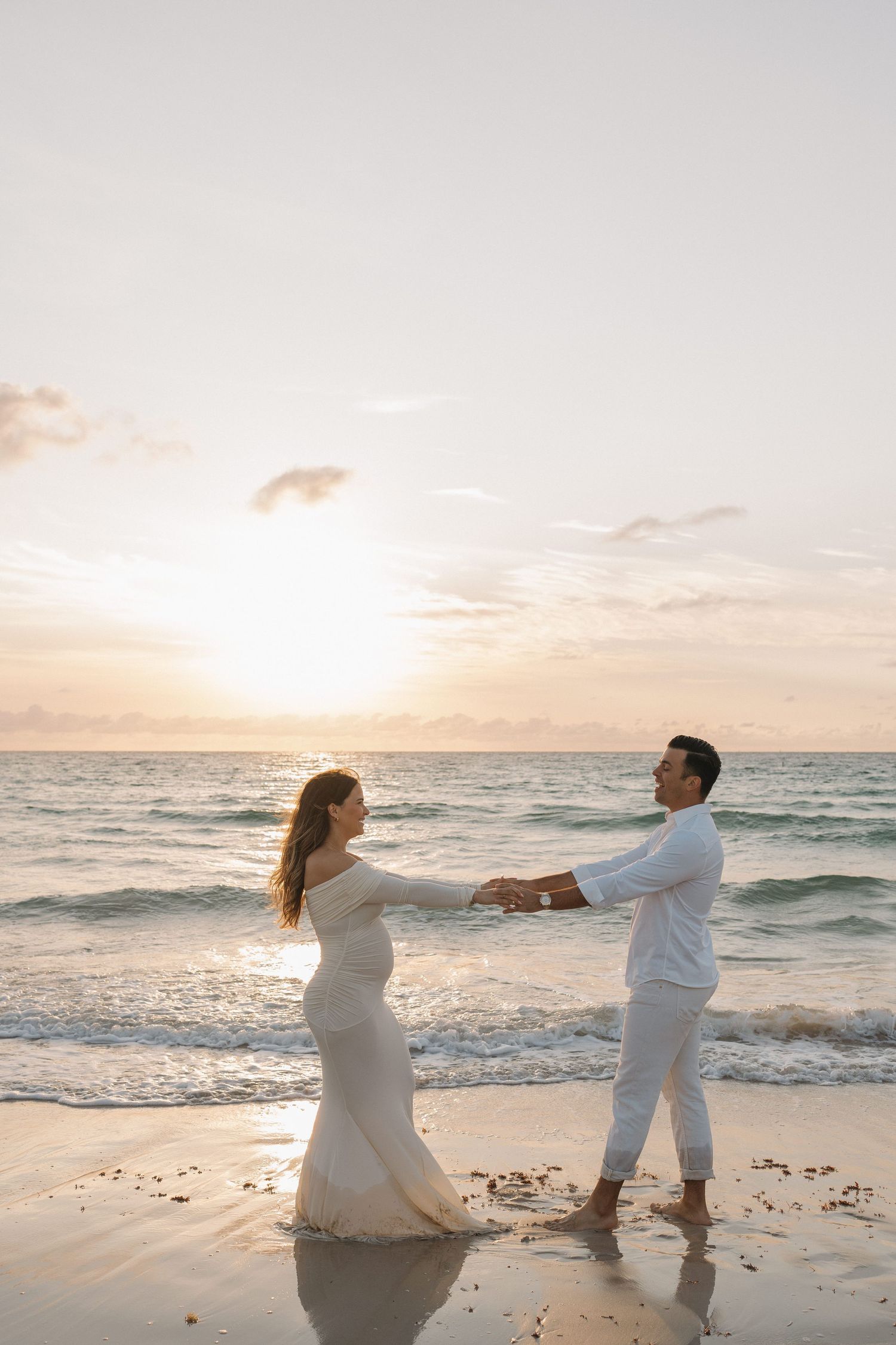 A romantic couple in white outfits dances together on a beach at sunset.