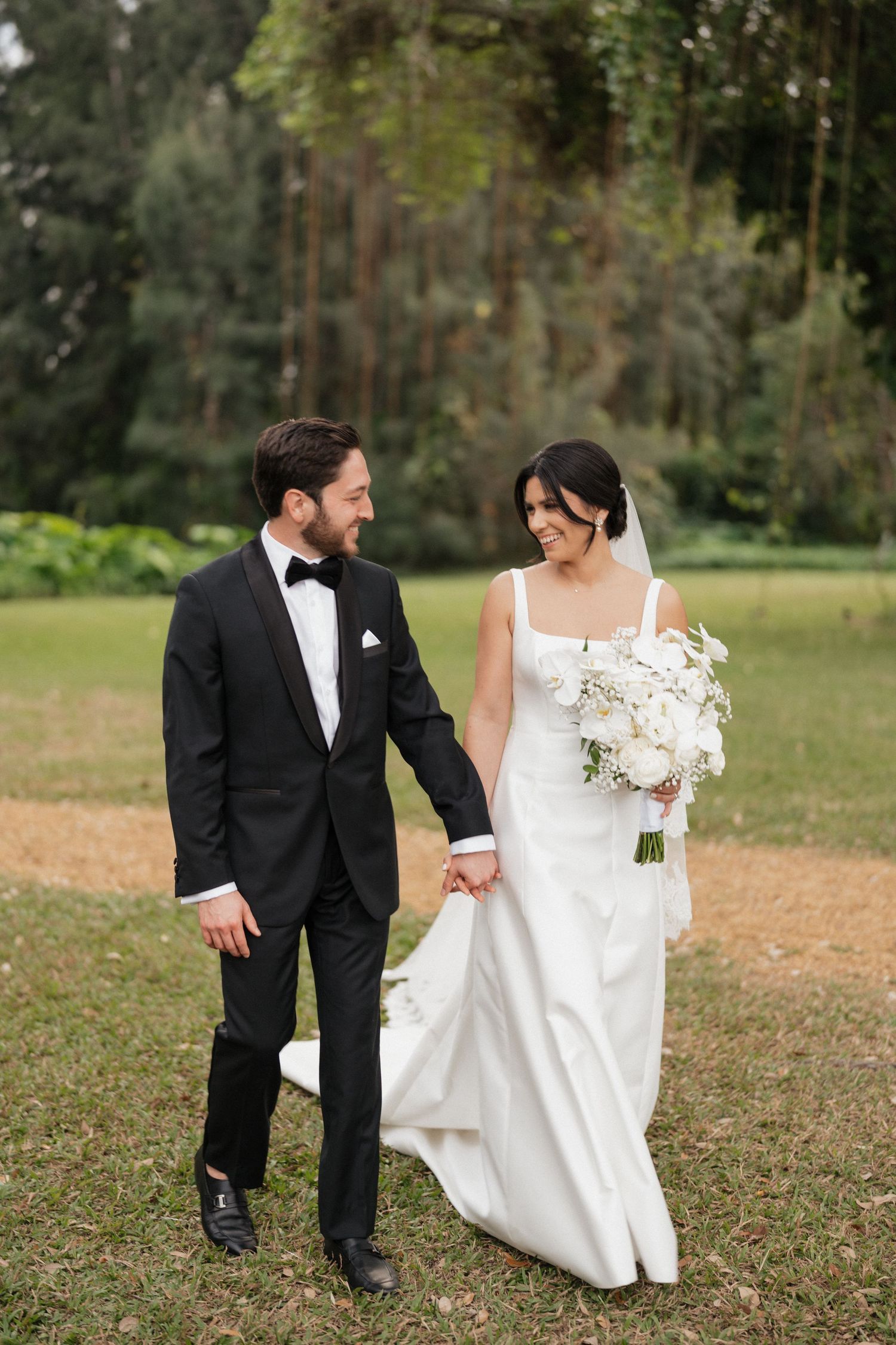 A wedding couple walks hand in hand on a grassy lawn with tall trees in the background.