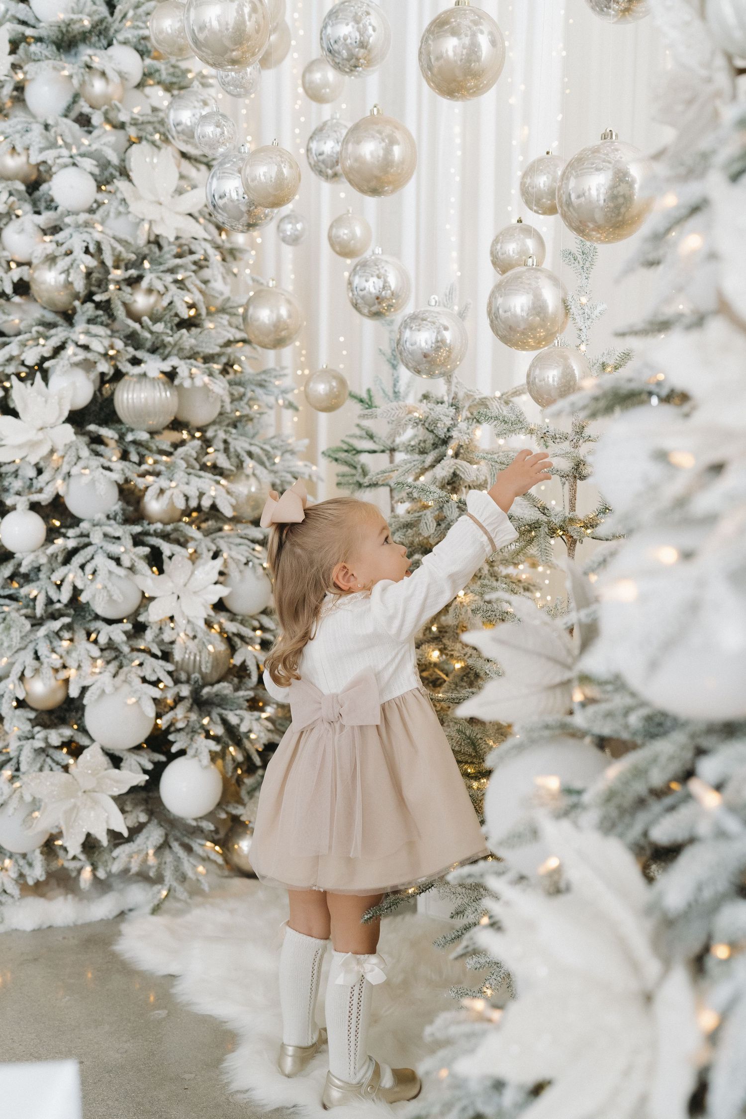 A little girl in a beige skirt reaches for silver Christmas ornaments on a decorated white tree.