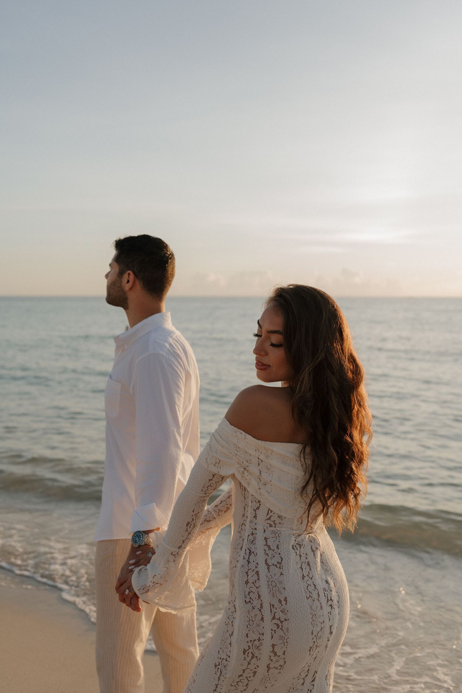 A couple in white clothing stands on the beach during golden hour with waves lapping at the shore.