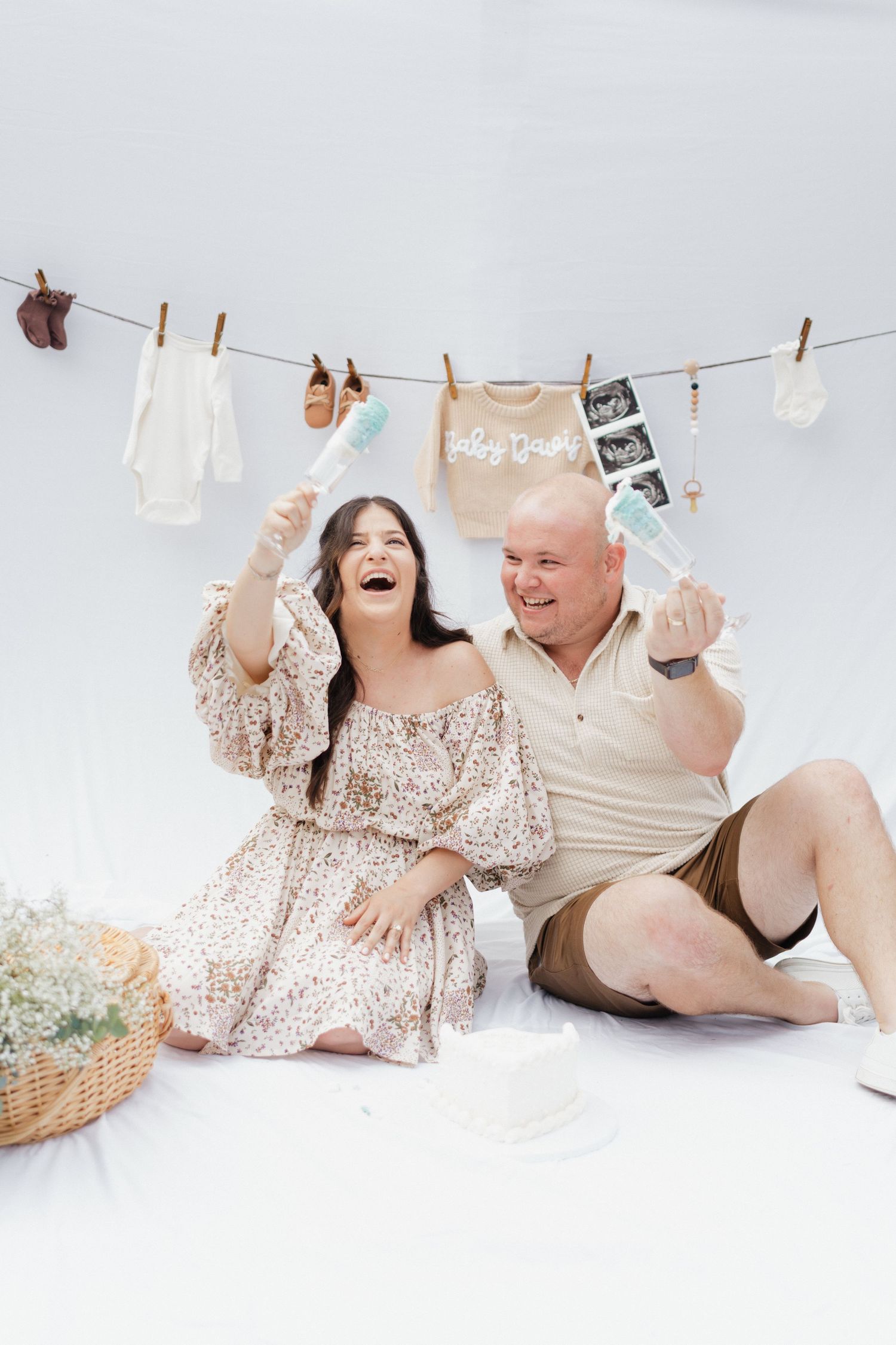 Pregnancy announcement photoshoot with baby clothes and ultrasound photos on clothesline against white backdrop.