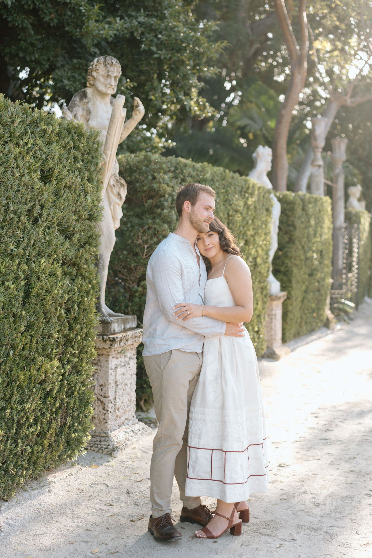Romantic garden scene with a couple embracing beside classical stone statues and manicured hedges.