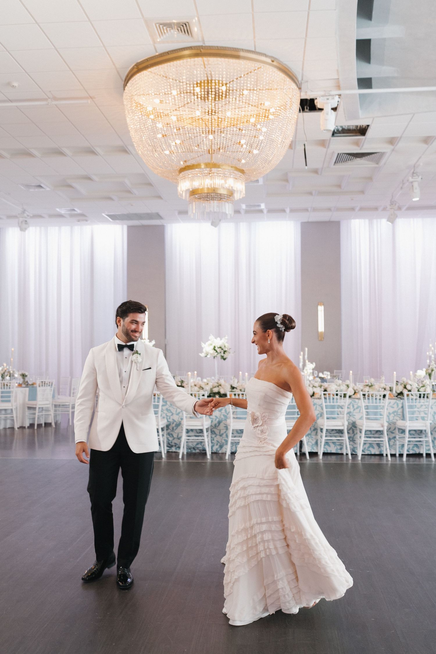 Wedding couple shares first dance under crystal chandelier in elegant reception hall.