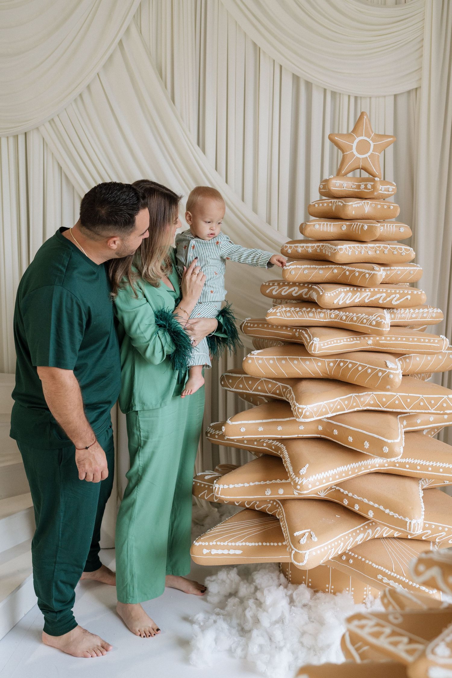 Family in green pajamas admires a gingerbread-style Christmas tree, with parents holding their baby near festive holiday decor.