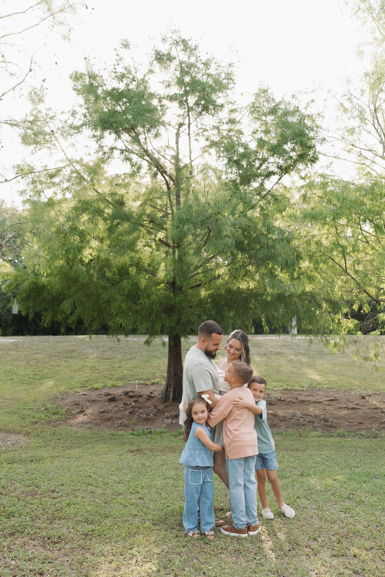 A family embrace under a leafy green tree in a peaceful park setting.