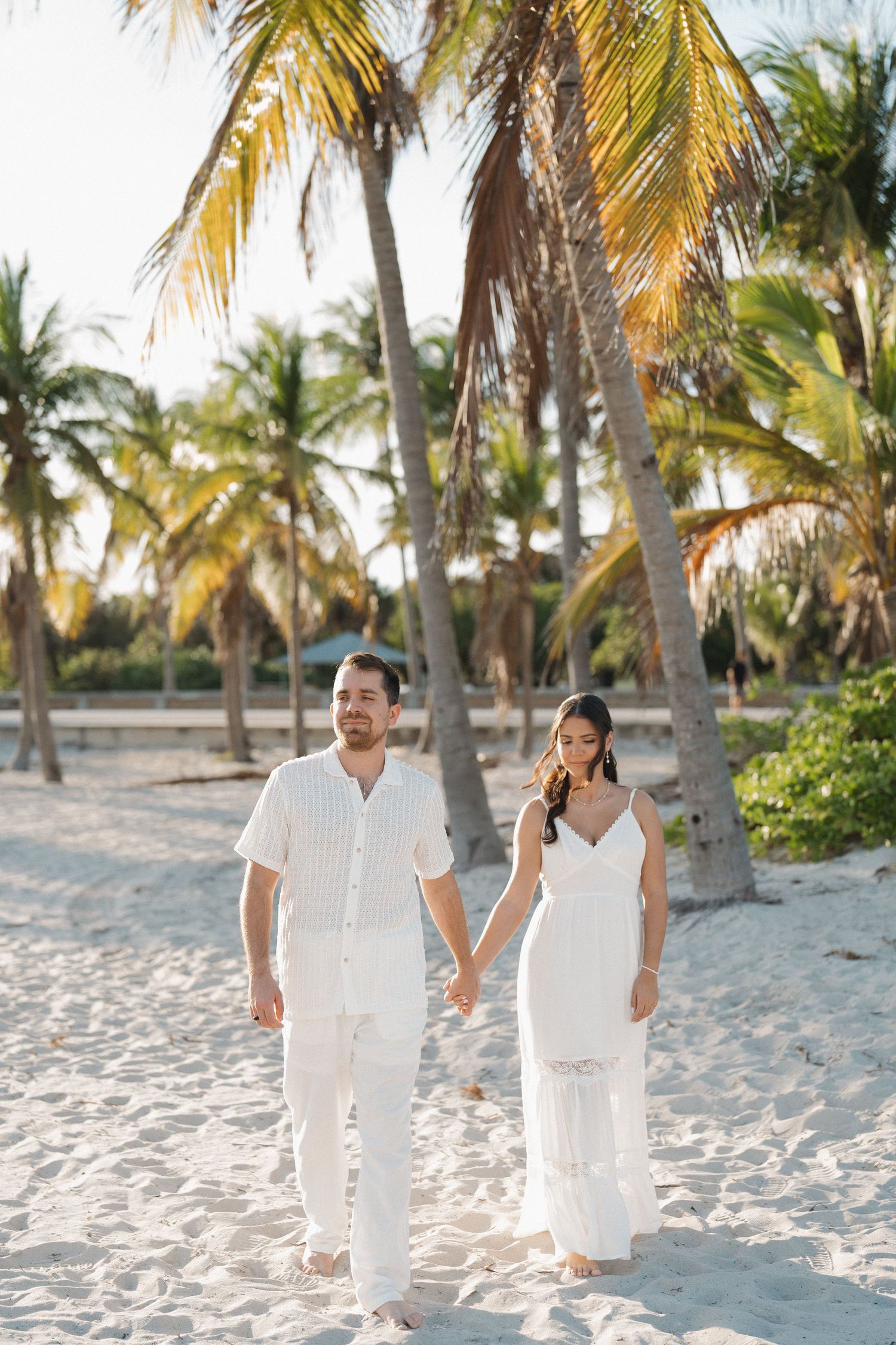 Romantic couple walking on a tropical beach with palm trees in all-white outfits.