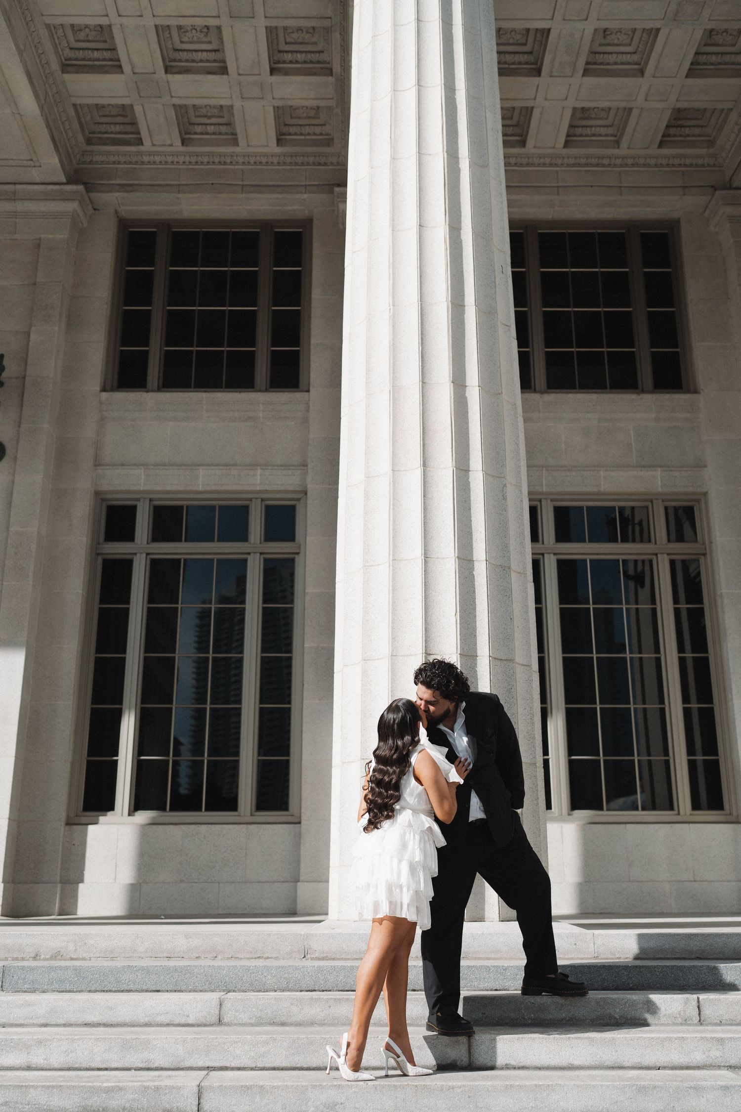 A couple shares a romantic moment against a white classical column on building steps.