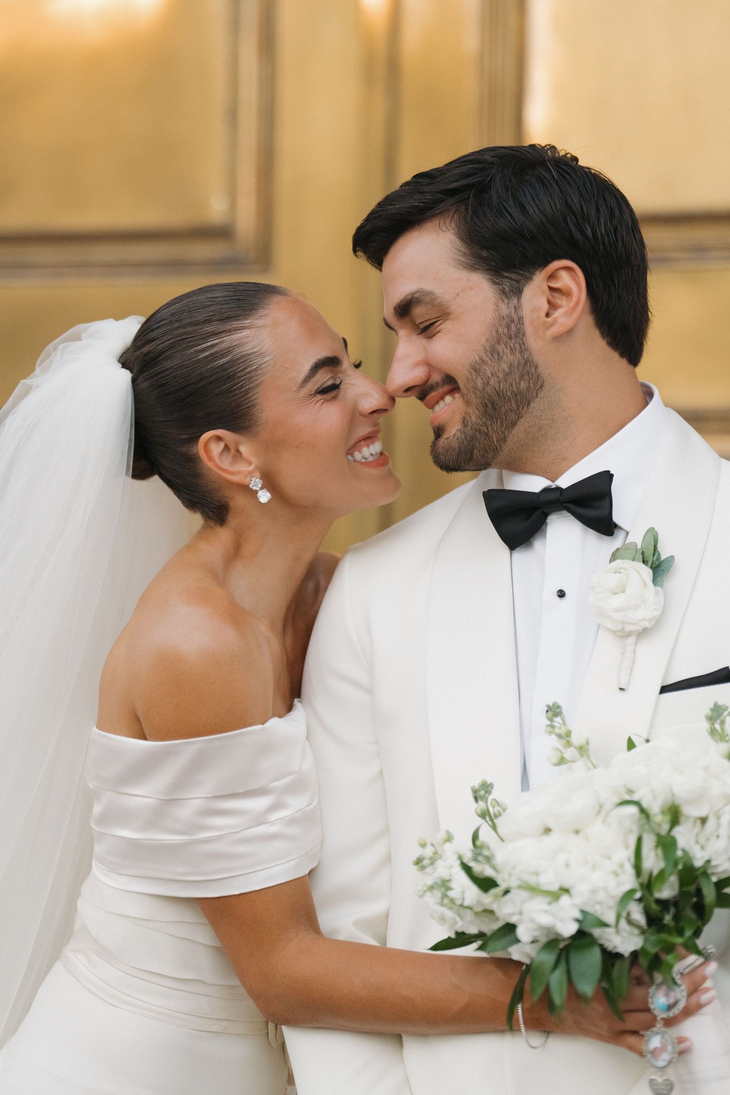 A bride and groom share an intimate moment while holding a white floral bouquet.