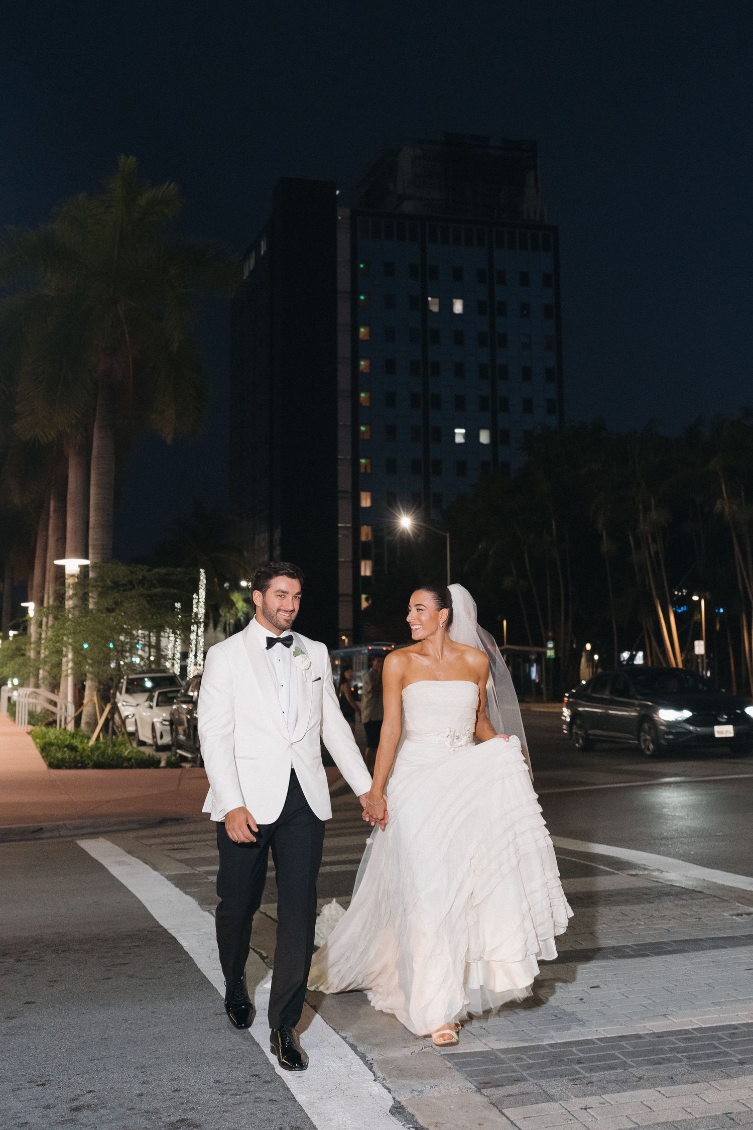 Newlyweds walk together at night along city street with palm trees and high-rise buildings.