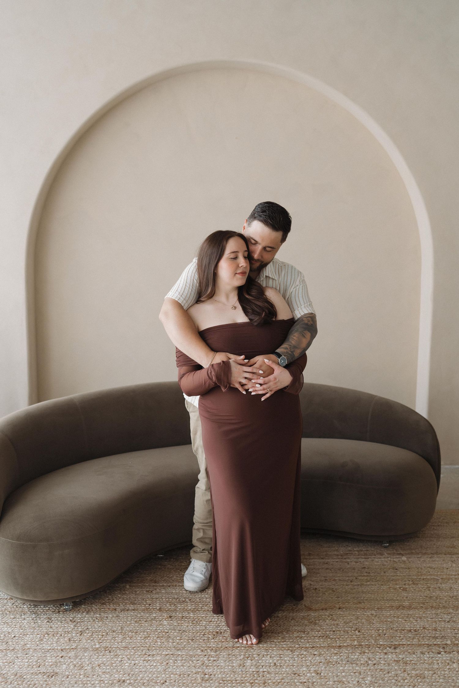 Intimate maternity portrait of couple embracing in front of curved archway and brown sofa.