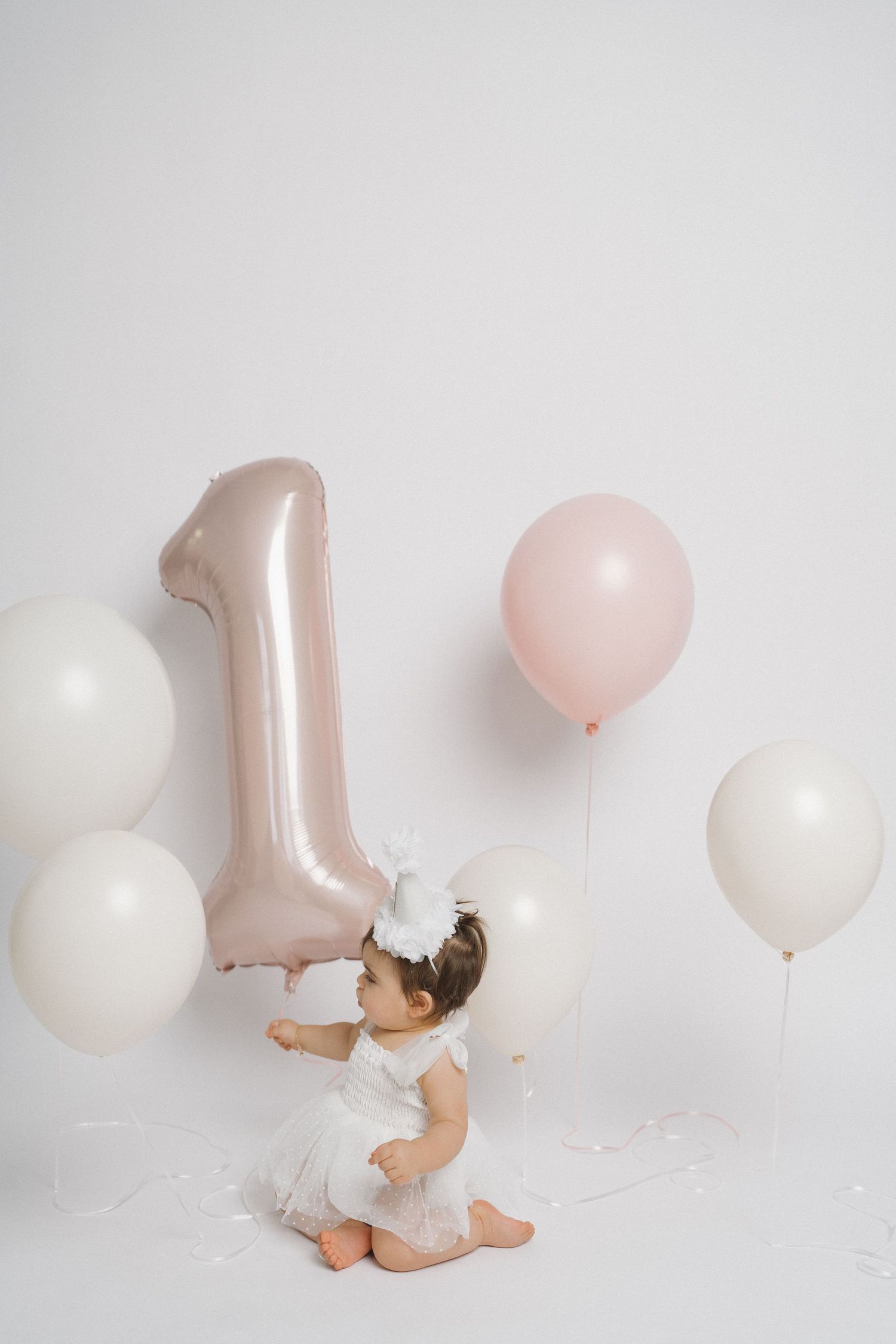 Baby in white dress crawls towards rose gold number one balloon and white balloons.