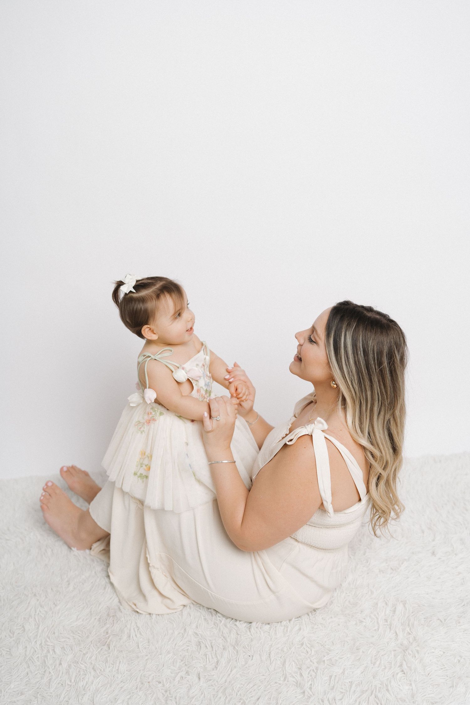 Mother and baby in matching cream outfits share playful moment on white carpet.