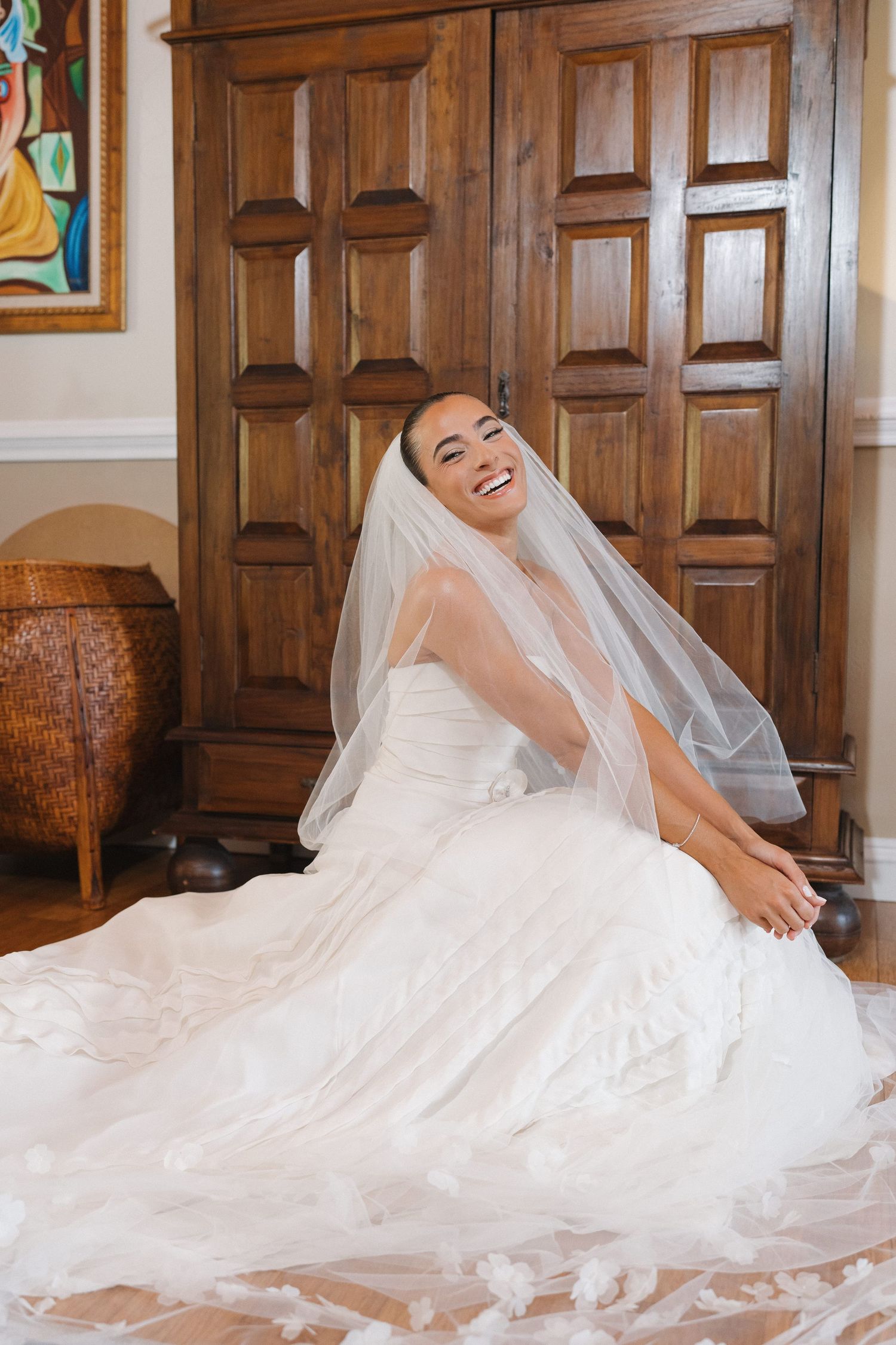 Bride in wedding dress and veil sits gracefully against wooden door.