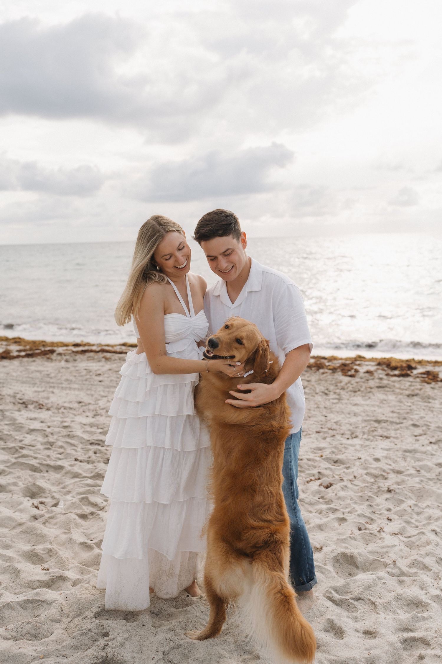 Couple laughing with excited golden retriever standing on hind legs at beach.