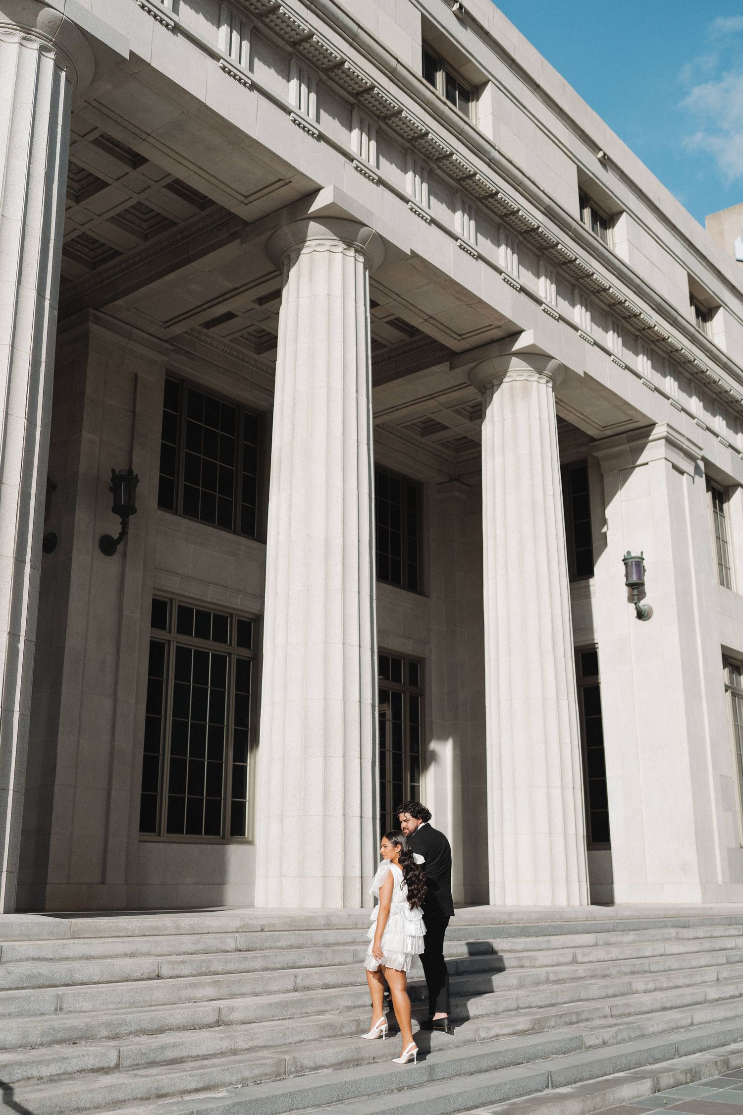 A couple stands on steps of a neoclassical building with towering white columns.