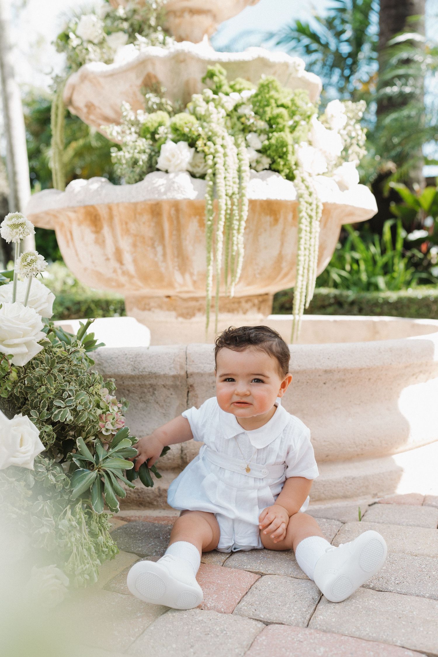 Baby sitting by ornate garden fountain surrounded by white floral arrangements.