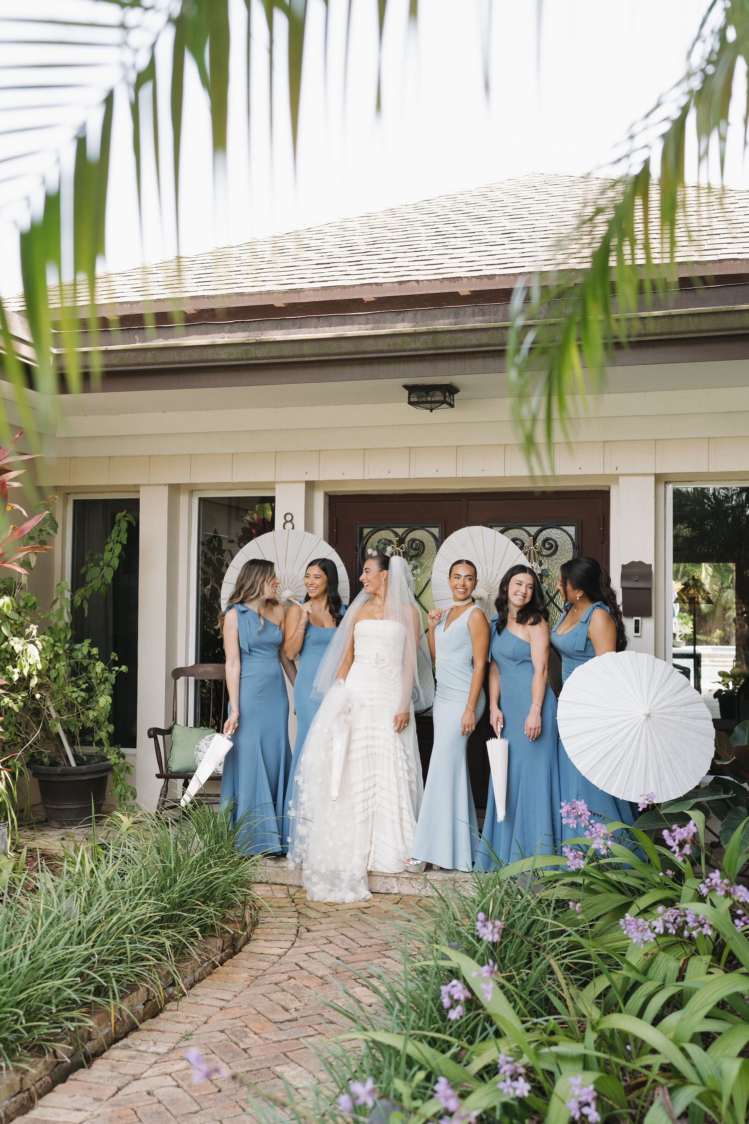Bride in white gown stands with bridesmaids in blue dresses on garden path.