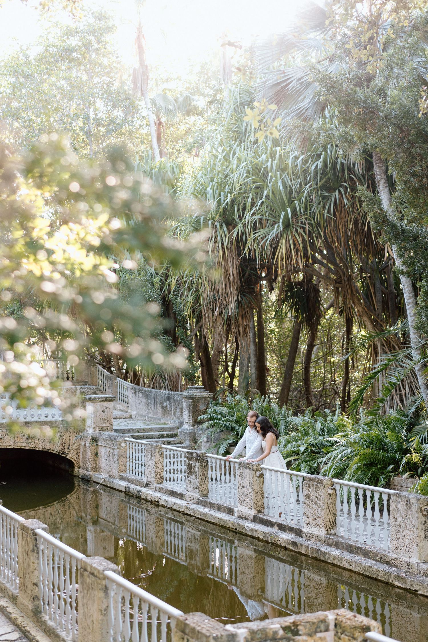 A couple stands on an ornate white bridge overlooking a pond in a lush tropical garden.
