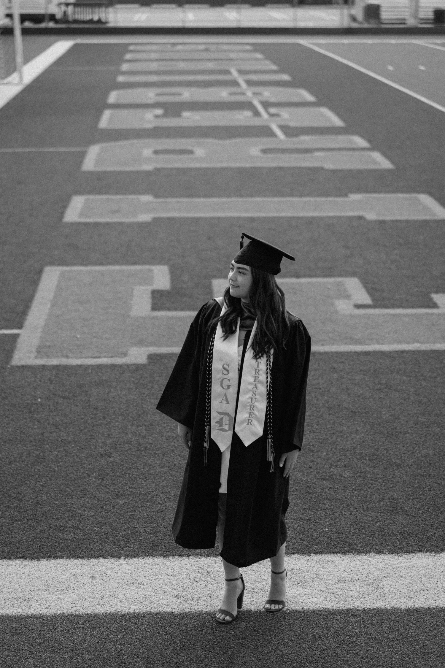 Graduate in cap and gown stands on football field in black and white photograph.