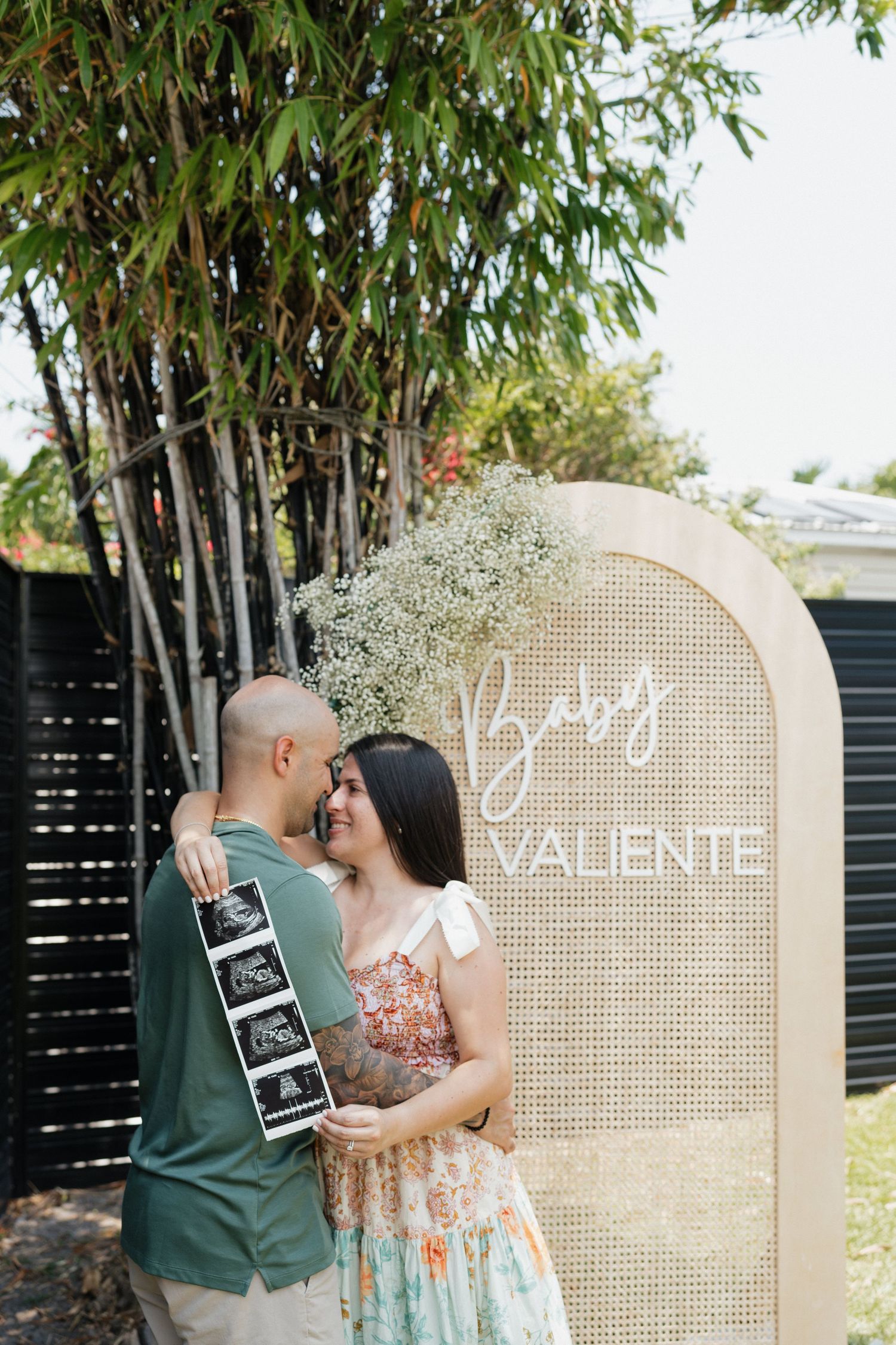 Couple poses with ultrasound photos in front of decorative beige arch at pregnancy announcement.