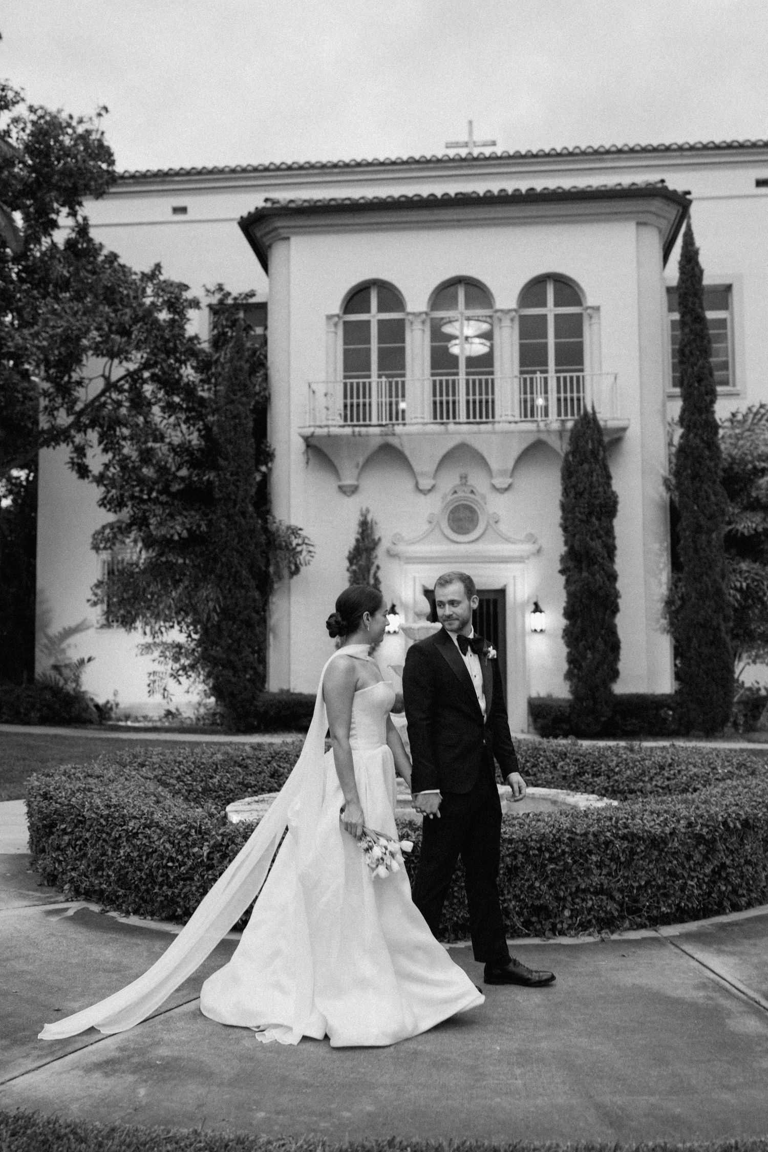 Classic black and white wedding portrait in front of Mediterranean villa with arched windows.