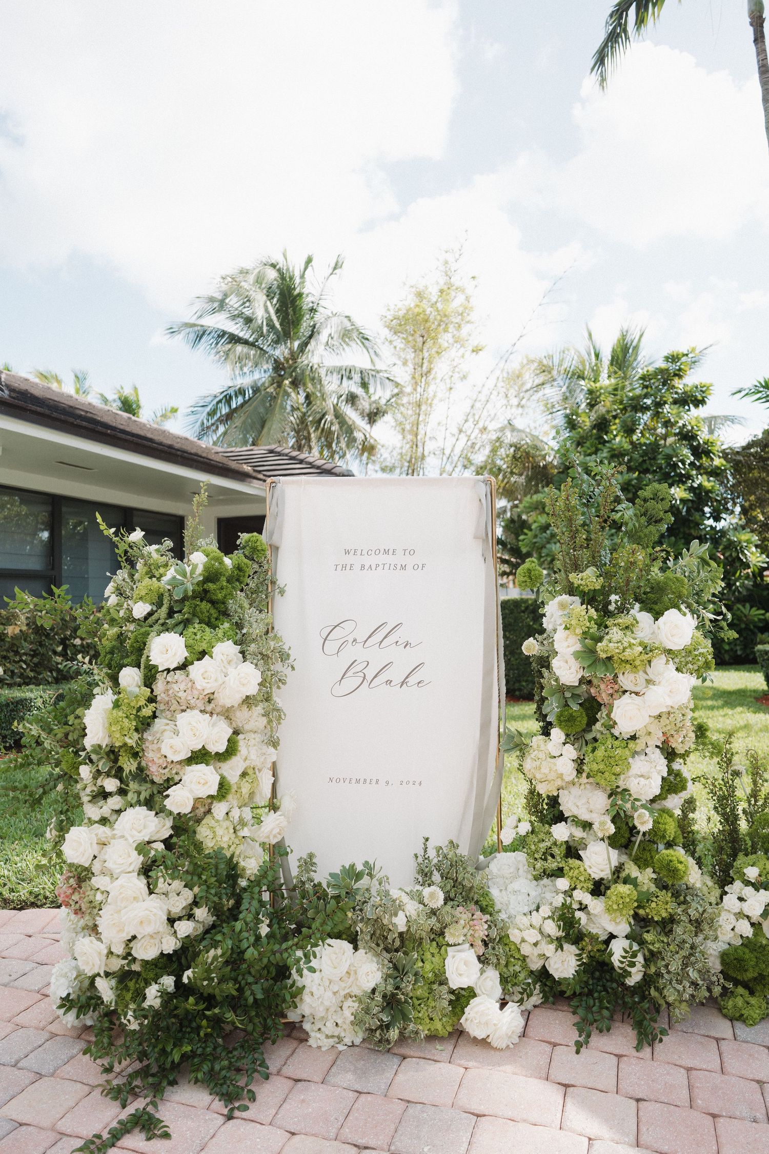White floral arrangements surrounding a wedding welcome sign outdoors with palm trees in the background.
