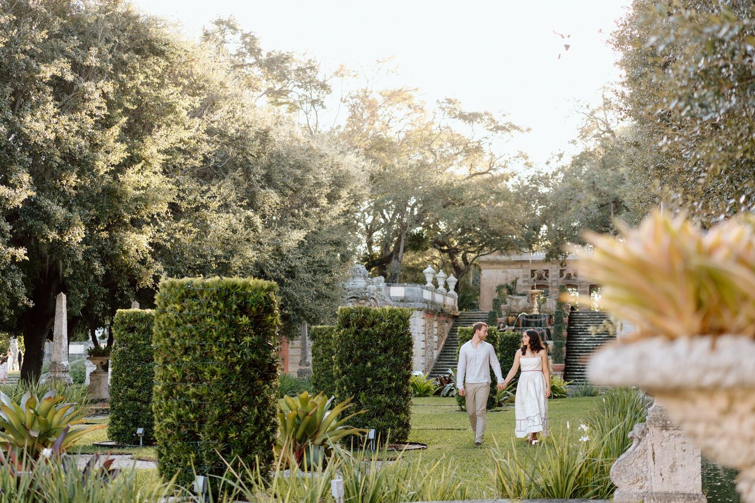 Couple walks hand in hand through a lush garden with manicured hedges at sunset.
