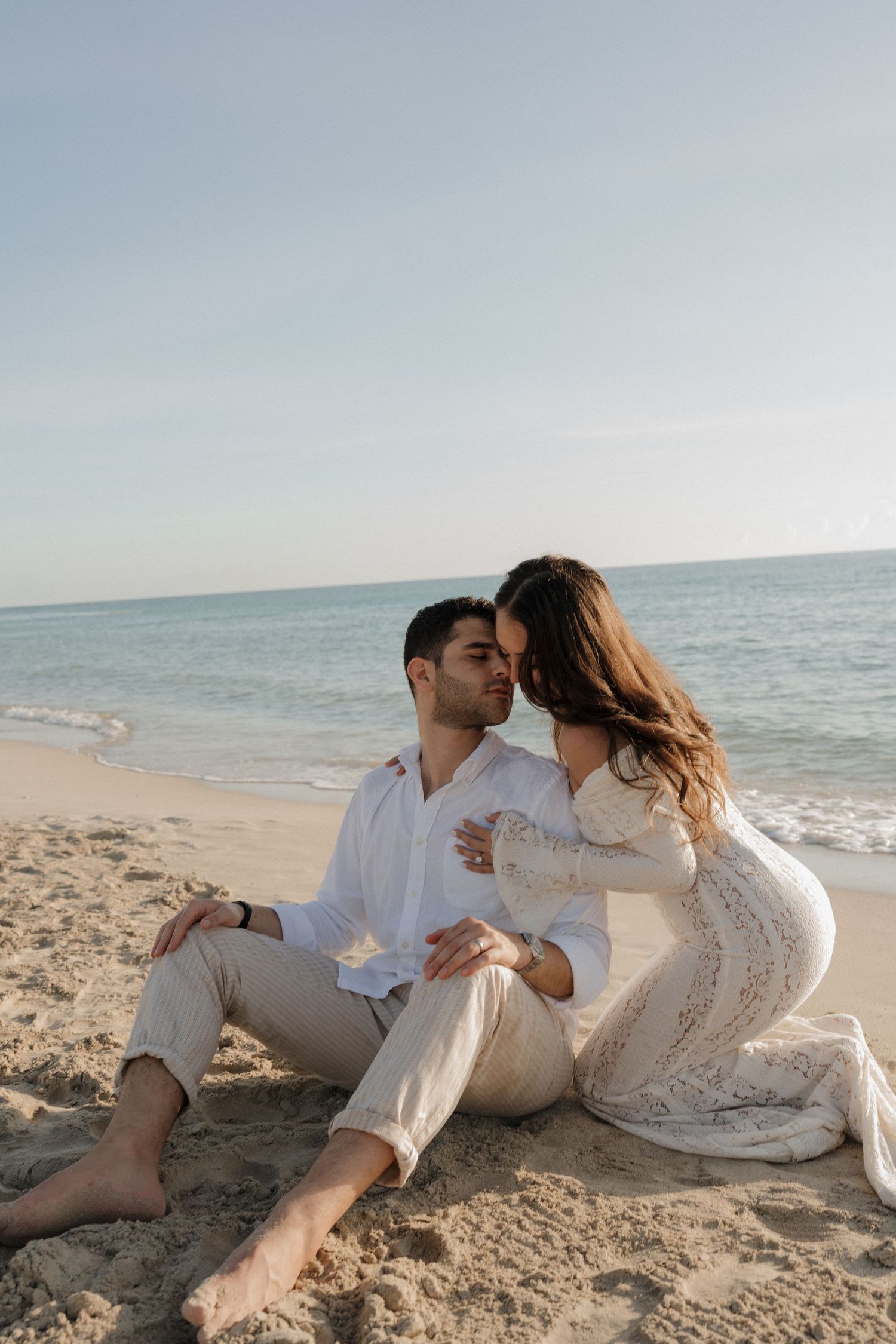 A couple sits intimately on the beach sand during a golden hour photoshoot.