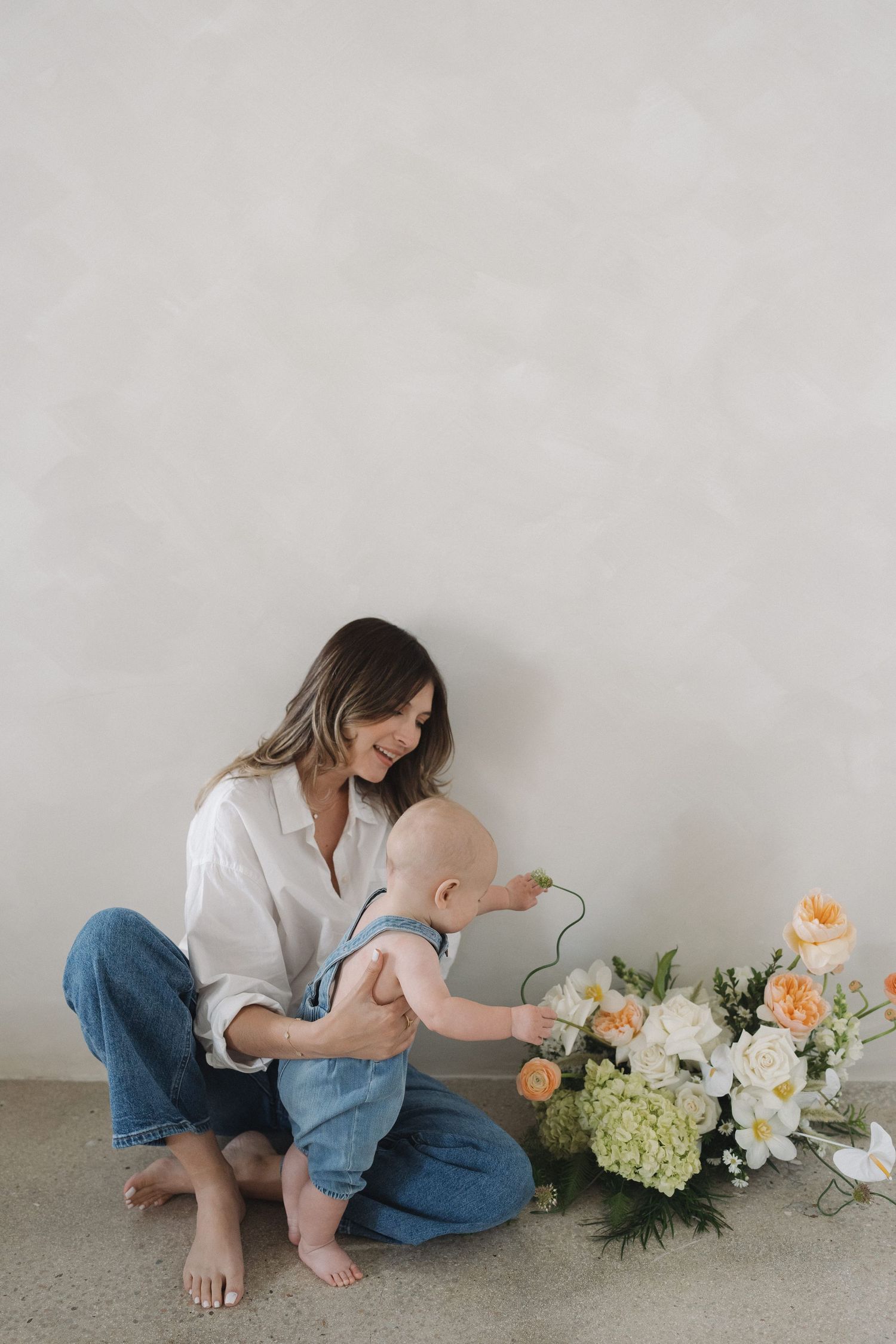 Mother and baby sit on floor by white wall with floral arrangement in minimal aesthetic photo series.
