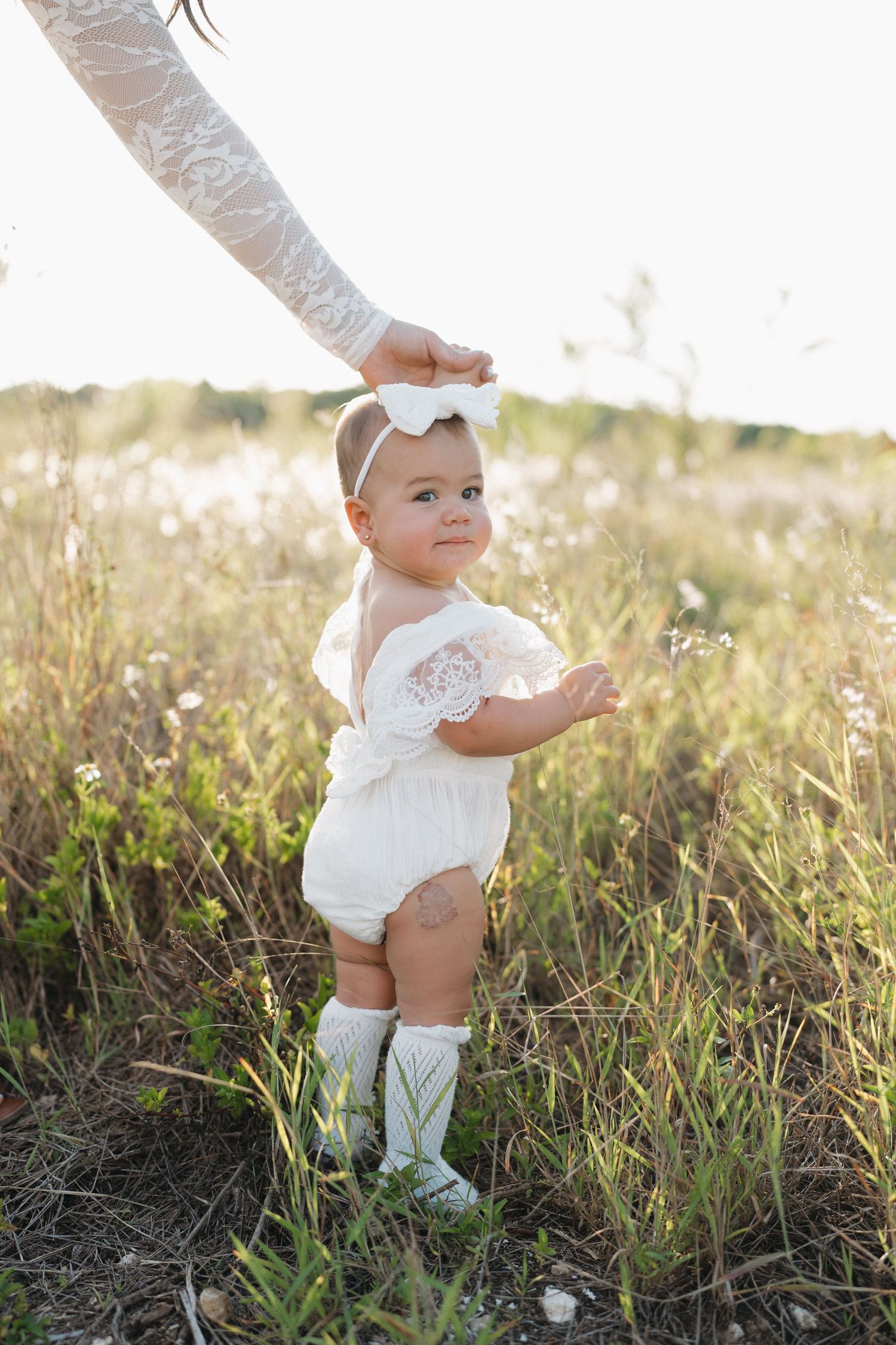 Baby in white romper and boots standing in sunlit wildflower field.