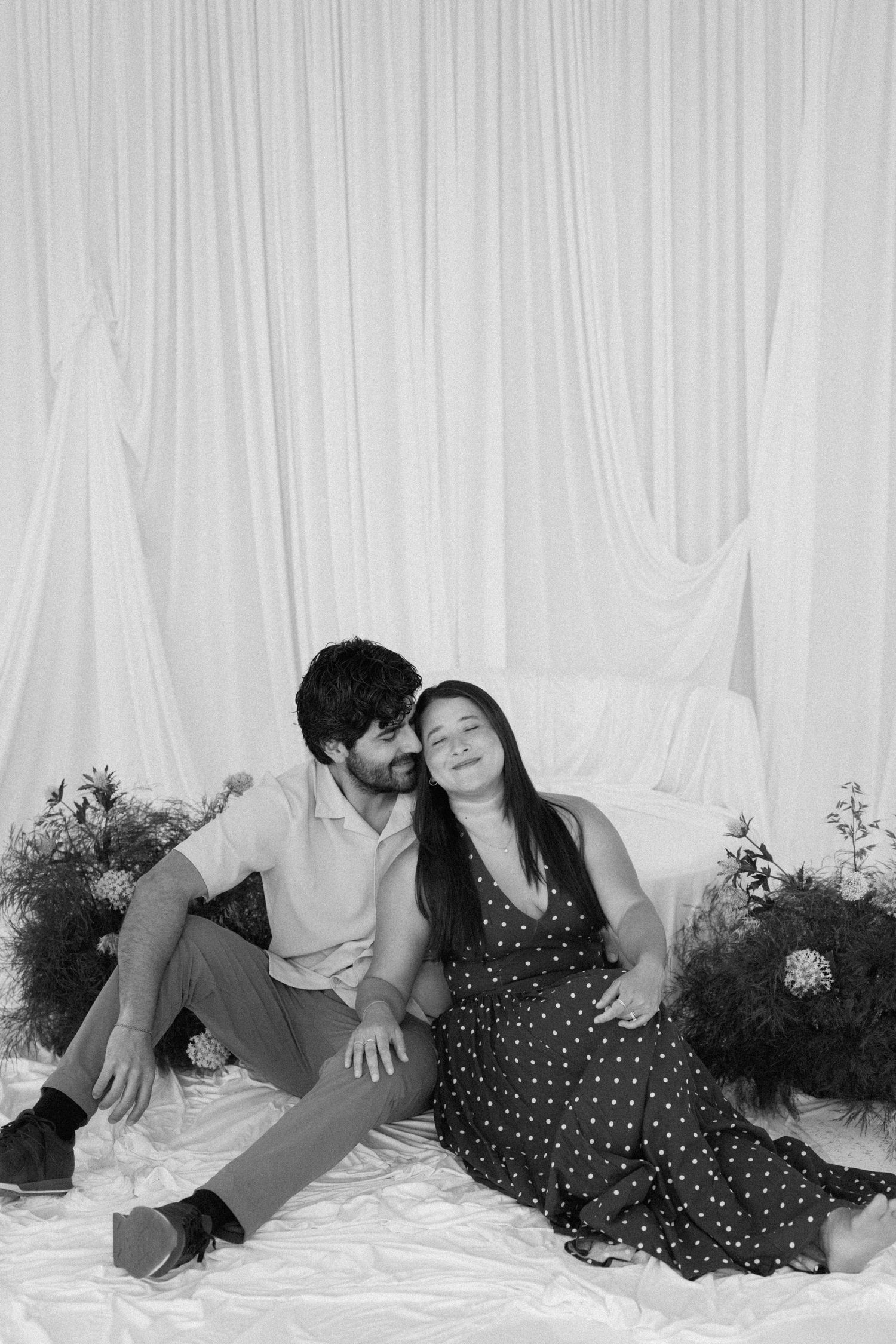 Black and white photo of a couple sitting together on white fabric, man kissing woman's cheek, surrounded by flowers.