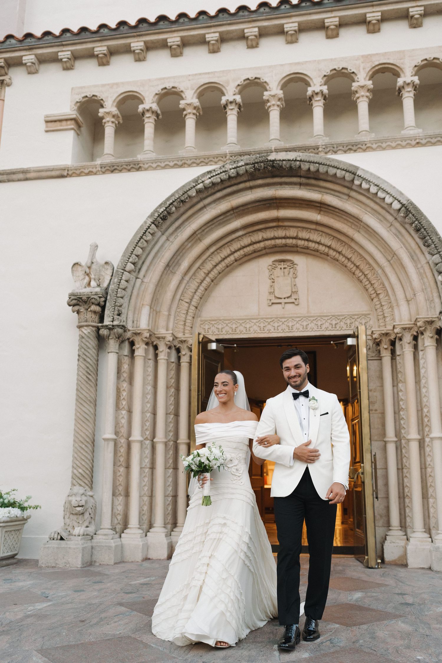 A wedding couple exits an ornate church with beautiful arched doorway and architectural details.