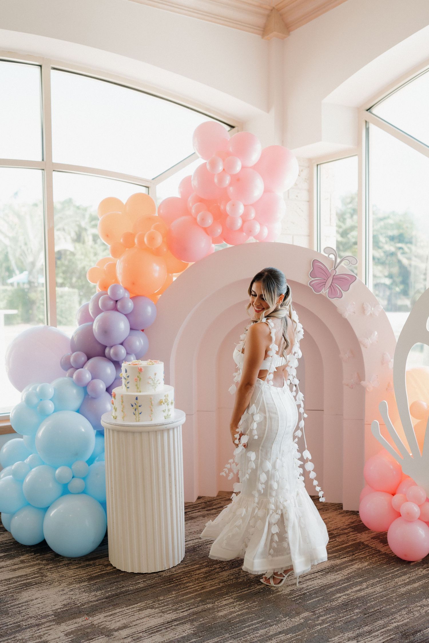 Pastel balloon arch with white column and butterfly decorations in sunlit room.