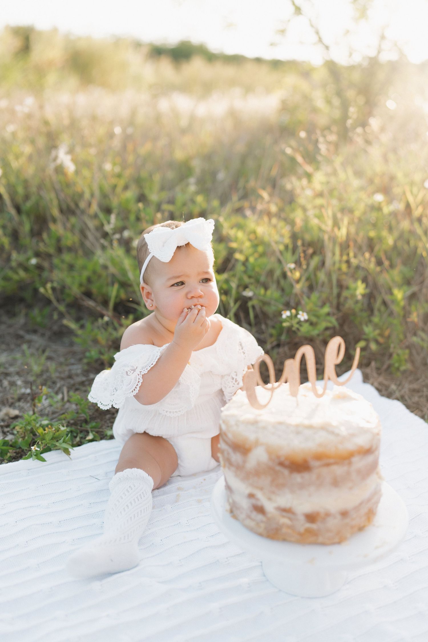 A baby in white enjoying their first birthday cake outdoors during golden hour.
