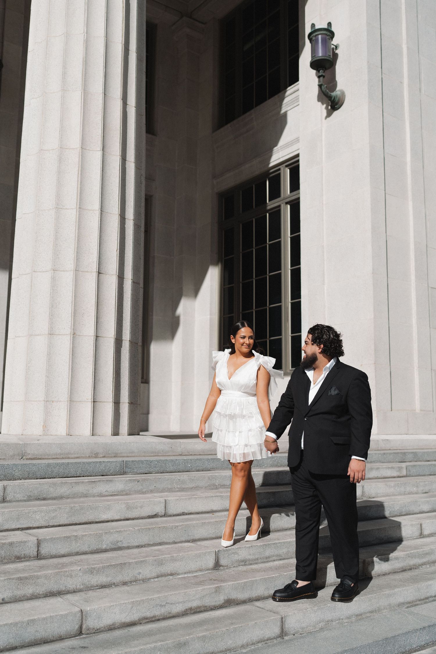 A couple in formal attire descends white marble steps of a classical building.