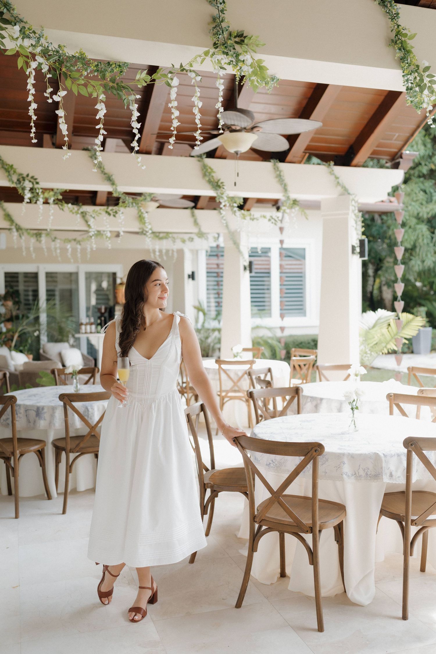 White dining area with cross-back chairs, hanging greenery, and string lights creating an elegant outdoor atmosphere.