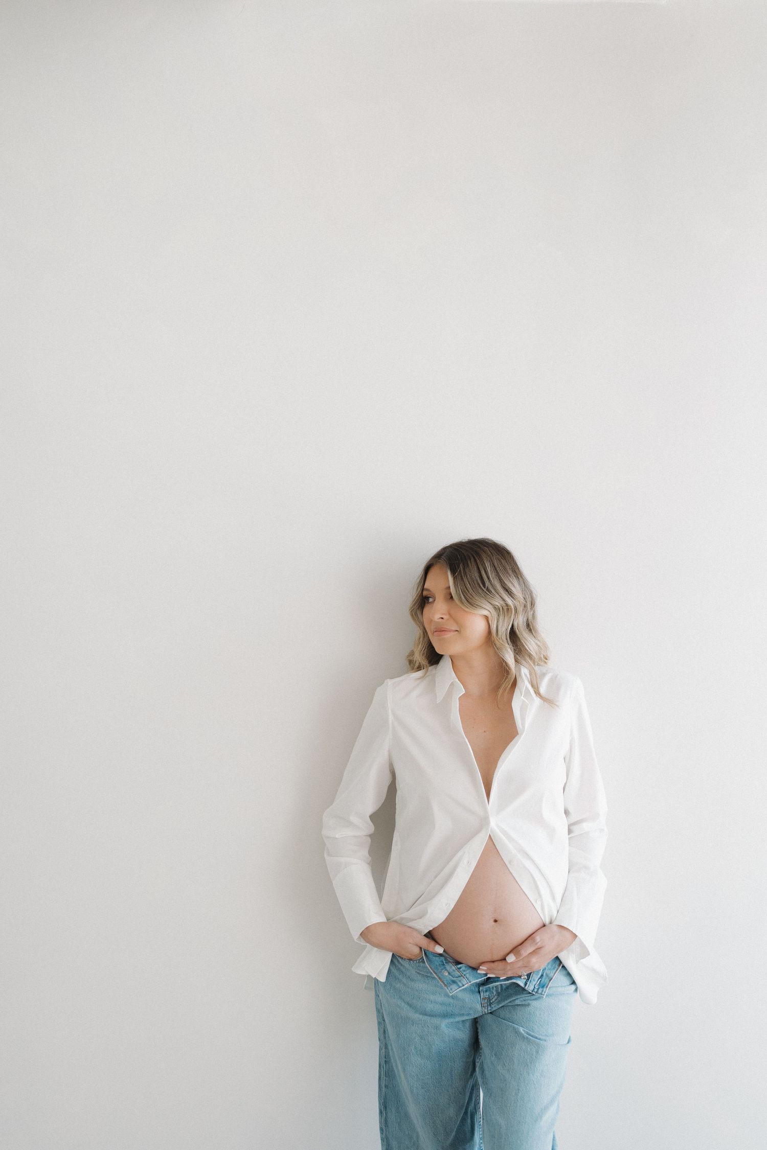 Candid maternity portrait featuring woman in white button-down shirt and jeans against white wall.