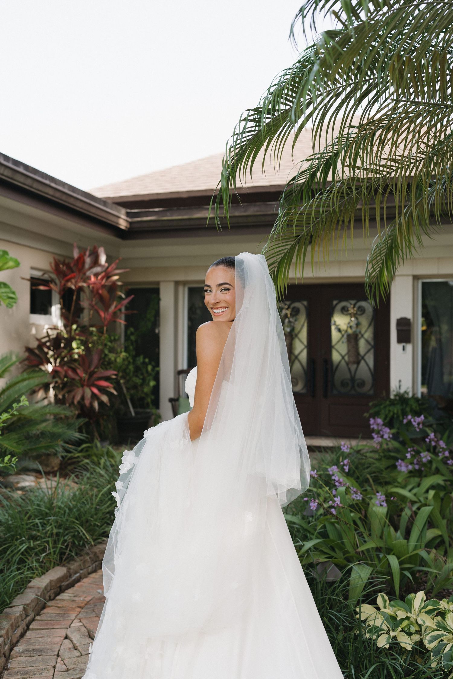 Bride in white gown smiles over shoulder in tropical garden setting.