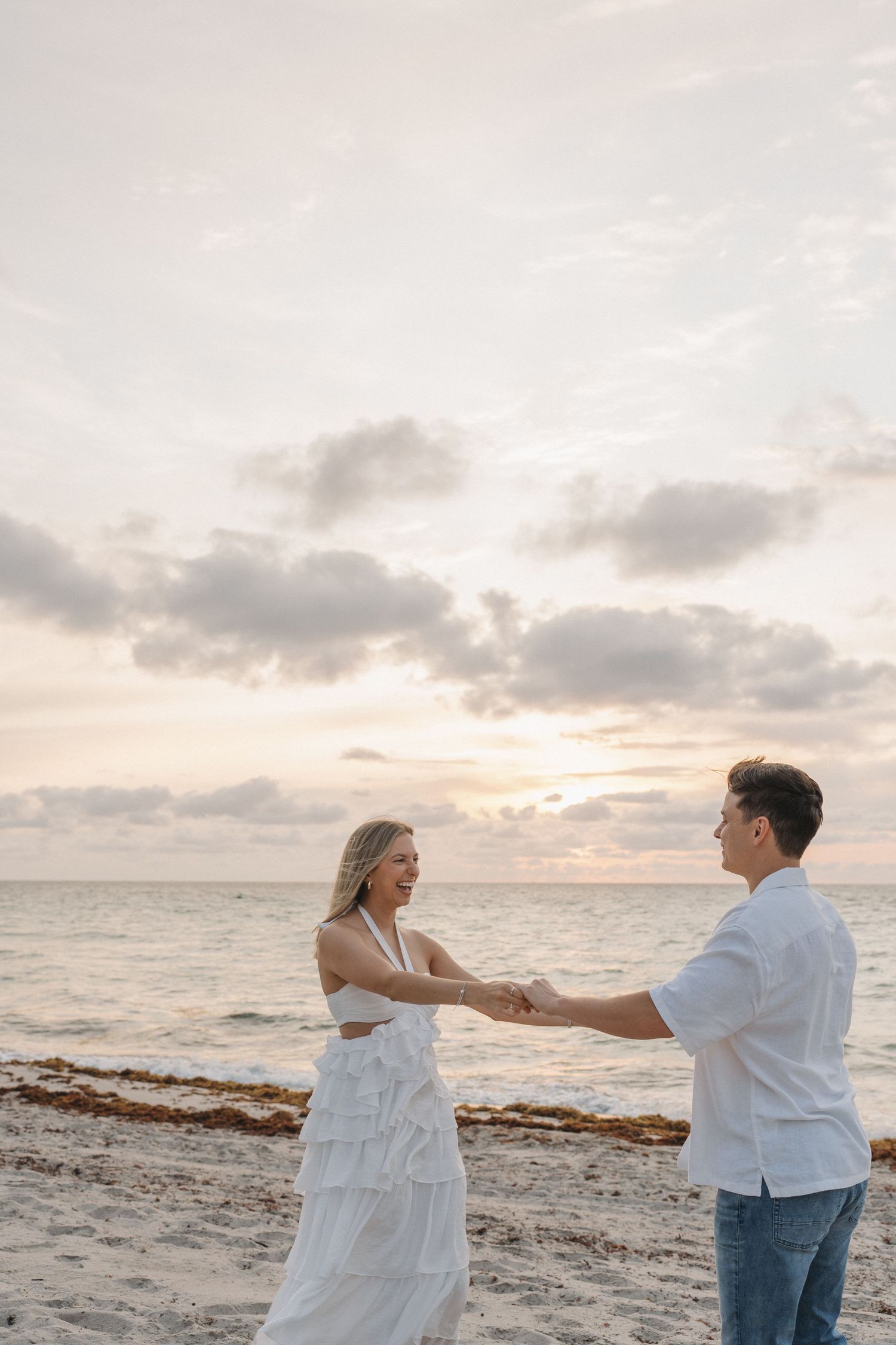 Couple dancing together on a beach at sunset with gentle waves and cloudy skies in the background.