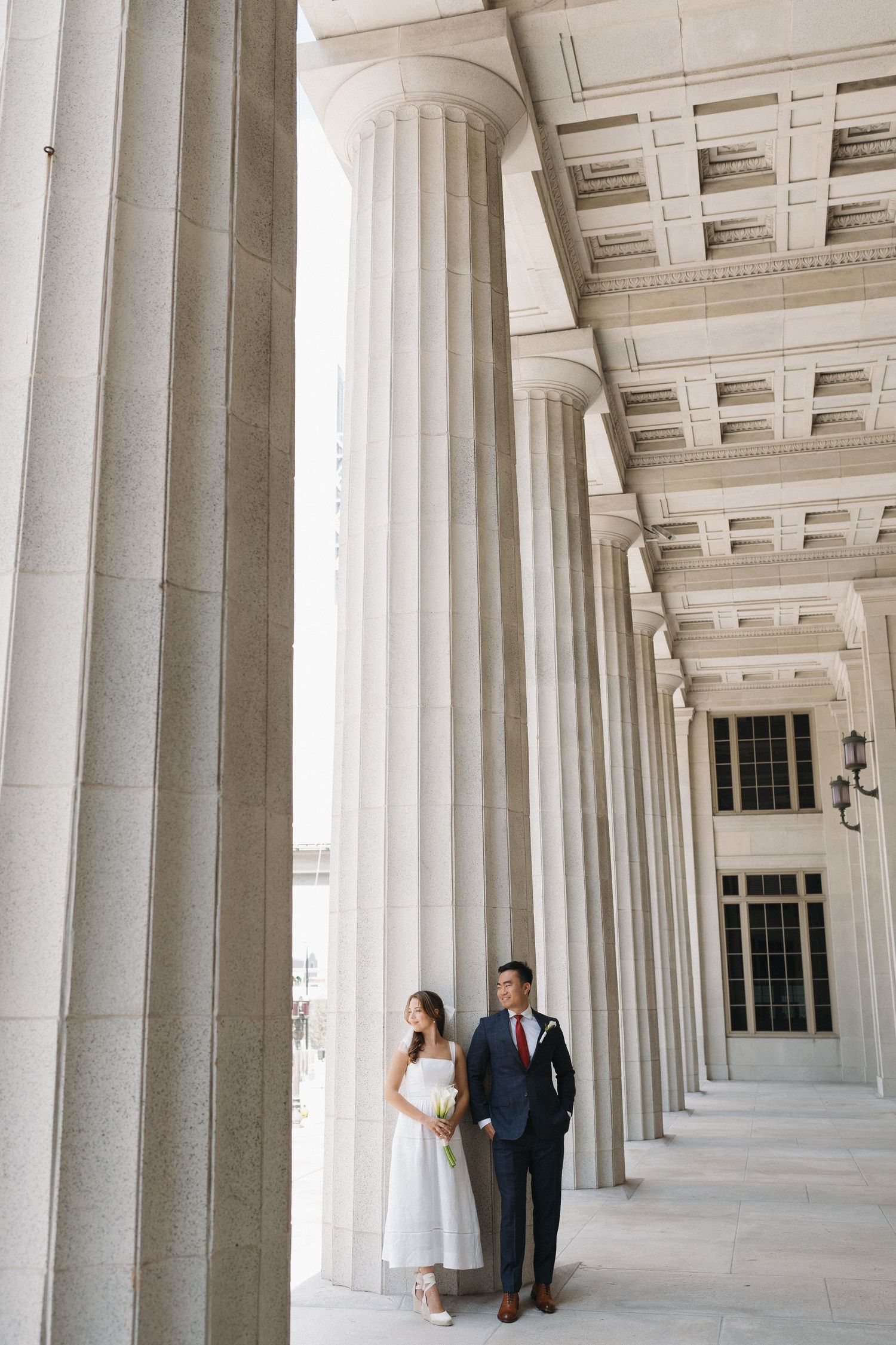 Couple poses between massive classical columns in formal wedding attire.