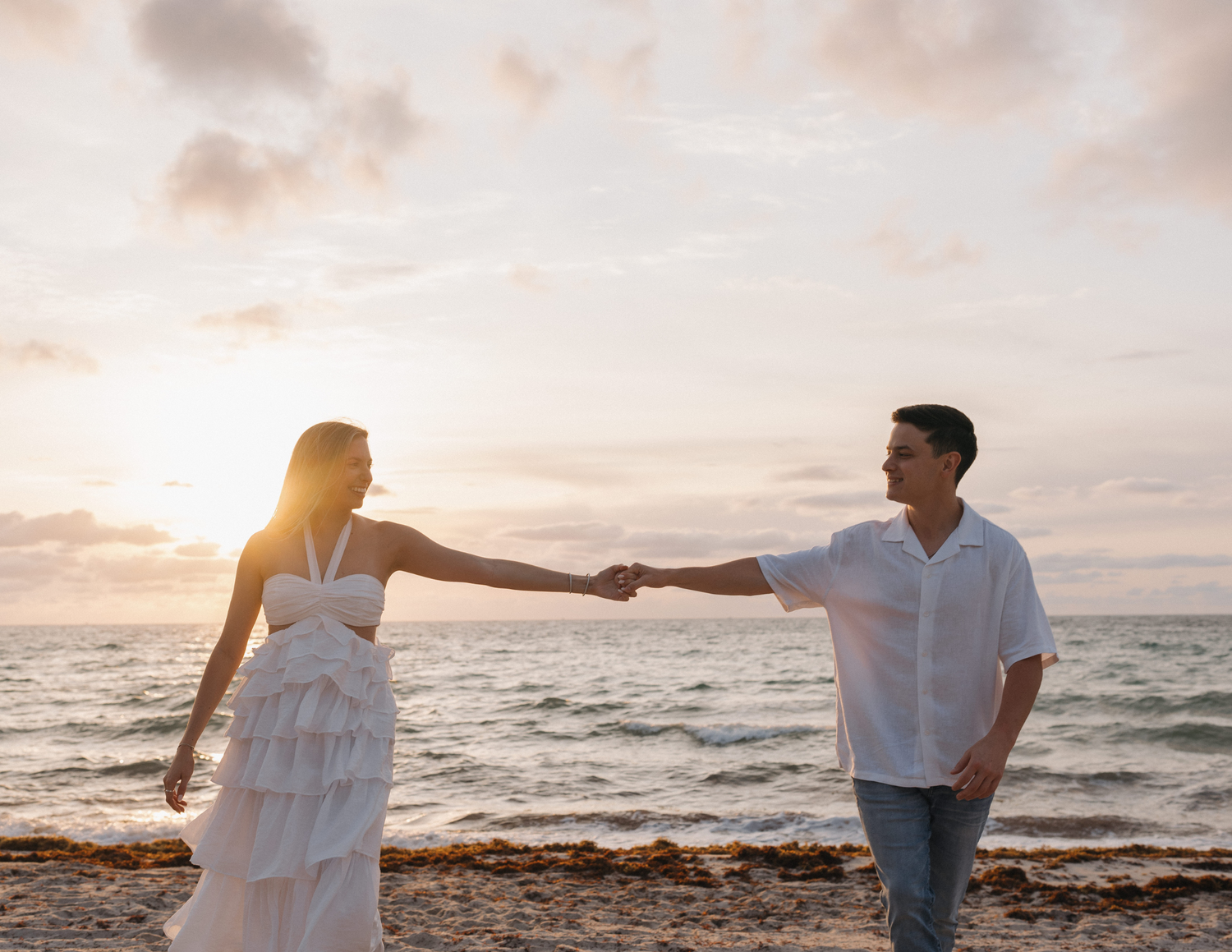Couple holding hands on a beach at sunset, woman in white ruffled dress, man in white shirt, ocean backdrop.