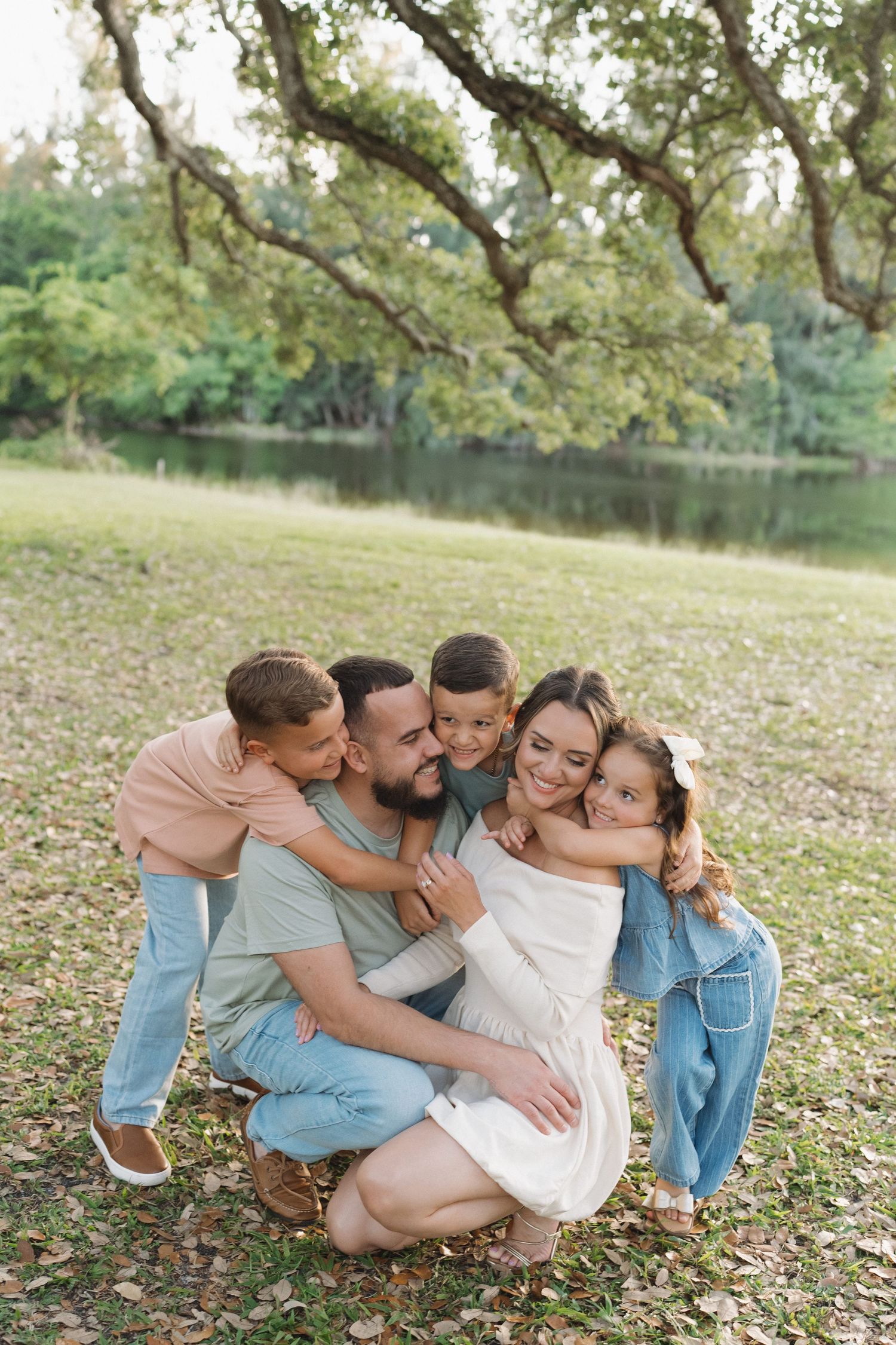 A heartwarming family group hug on grass under oak trees near a lake.