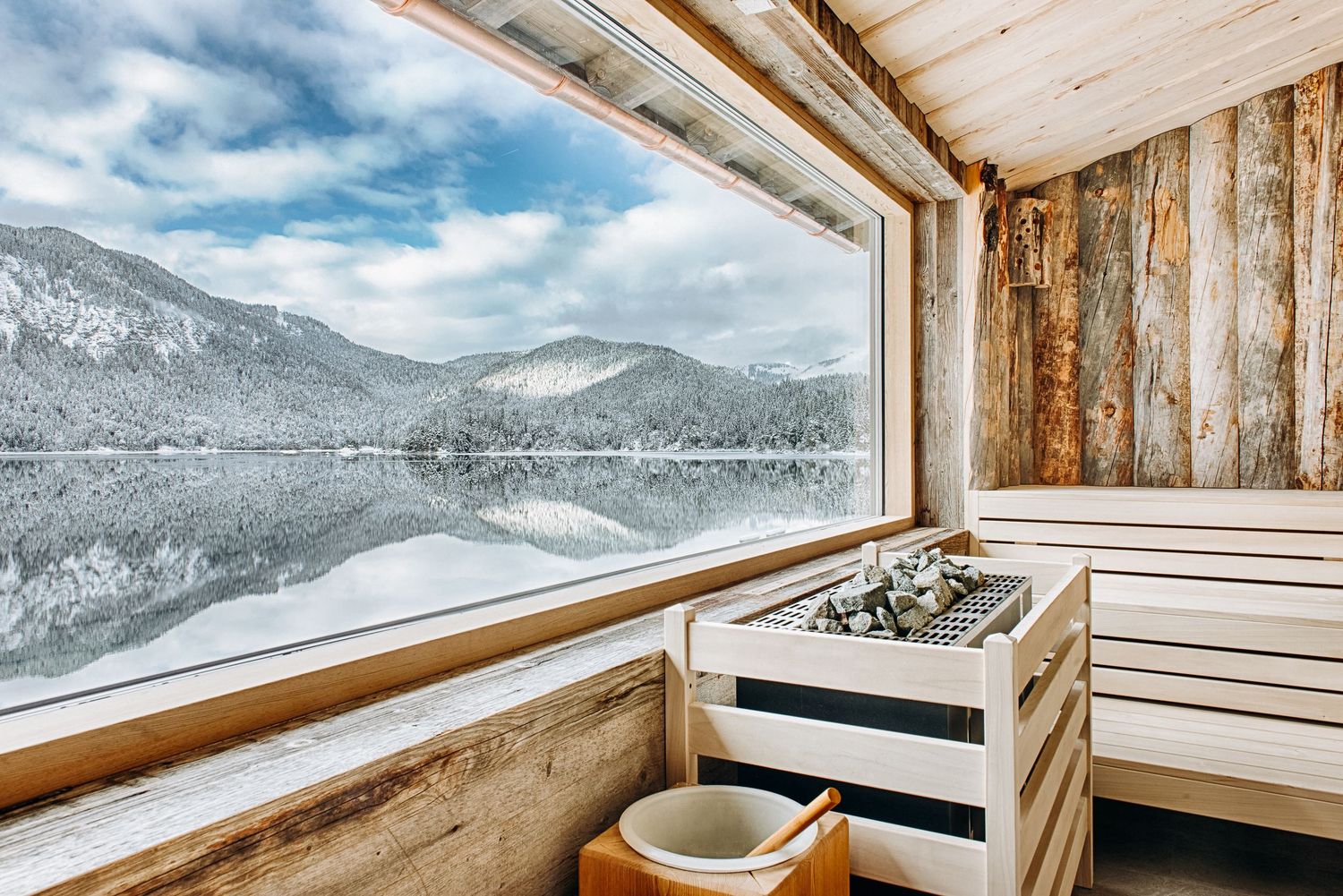 Luxurious sauna with panoramic window overlooking snow-capped mountains and pristine lake in winter.
