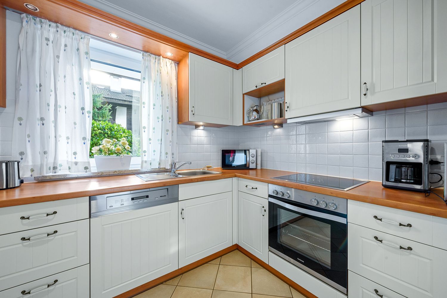 Modern white kitchen with wood trim features shaker cabinets, built-in appliances, and a bright window with sheer curtains.