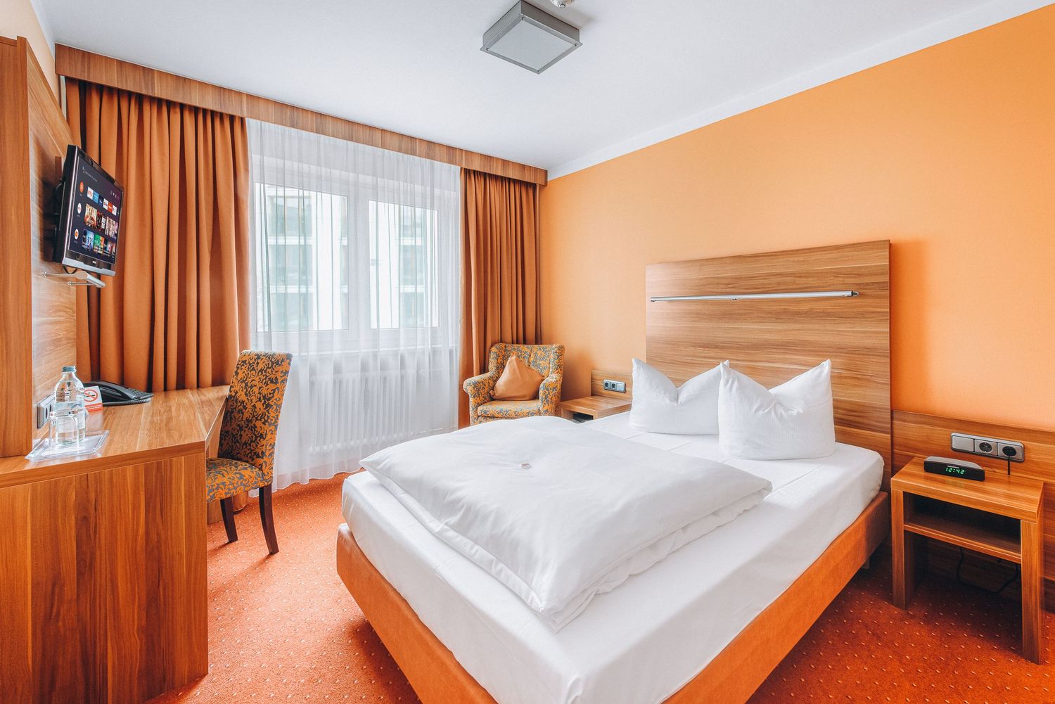Hotel room featuring orange walls, wooden furniture, and white bedding with matching curtains.