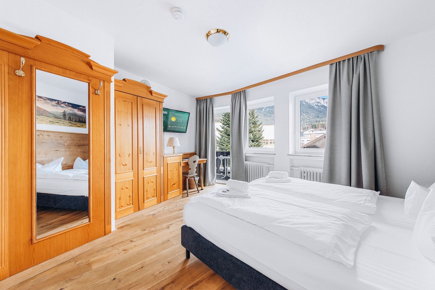 Bright hotel bedroom with wooden wardrobe, white bedding, hardwood floors and mountain views through the window.